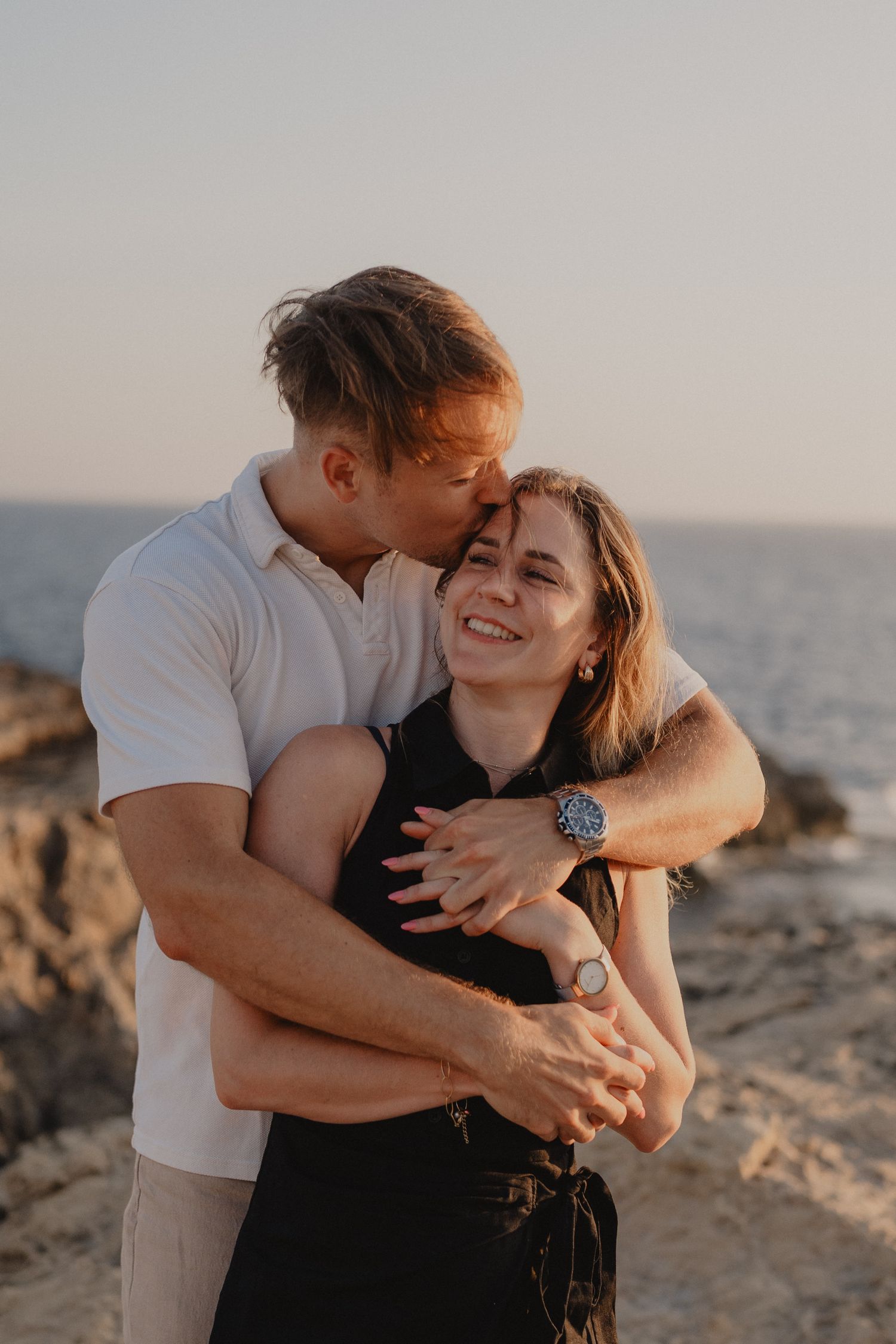 A couple embraces at sunset on rocky coastal cliffs, man kissing woman's temple while she smiles joyfully.