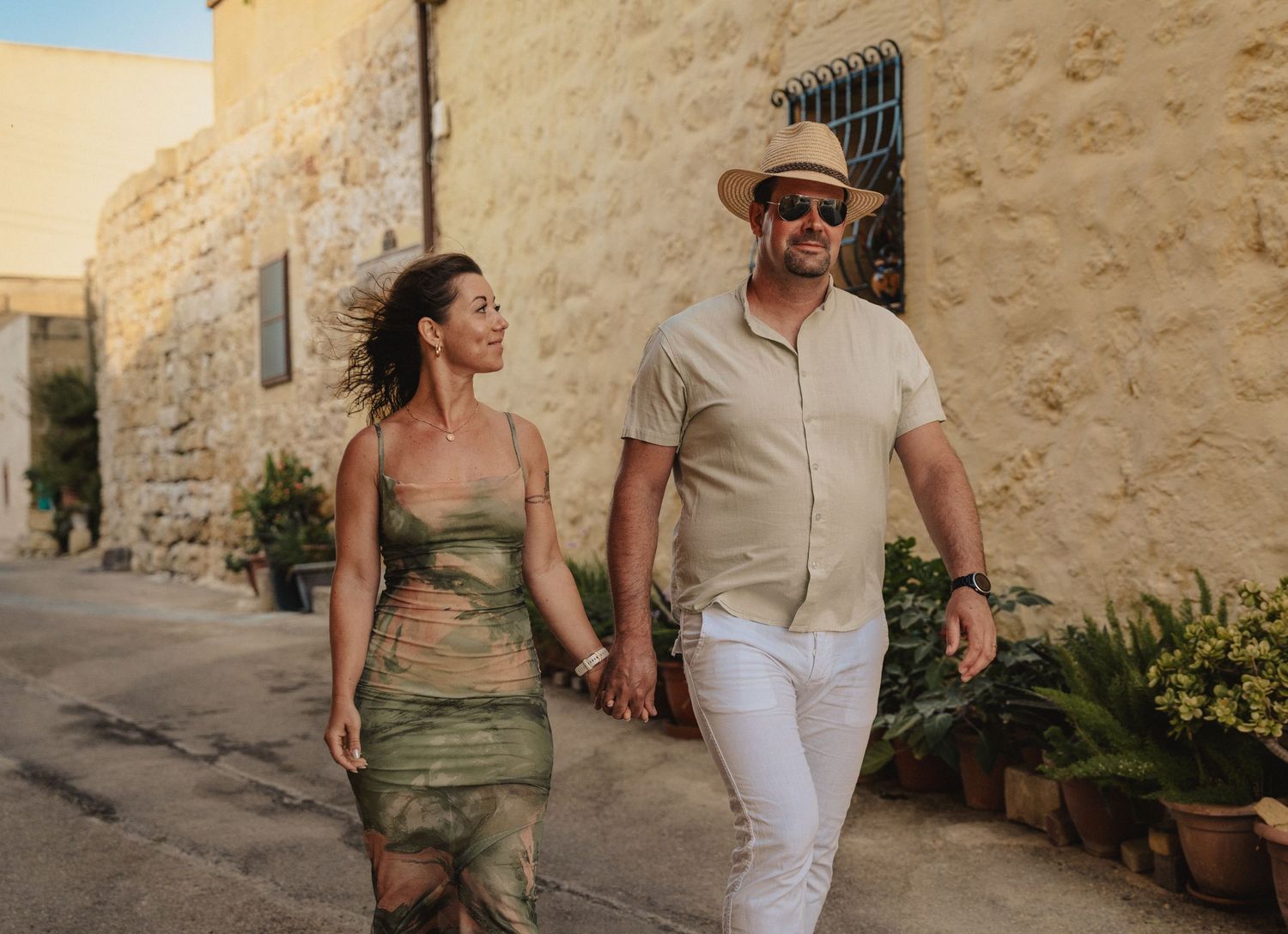 Couple holding hands walking through a sunny Mediterranean stone village street, woman in green dress, man in hat.