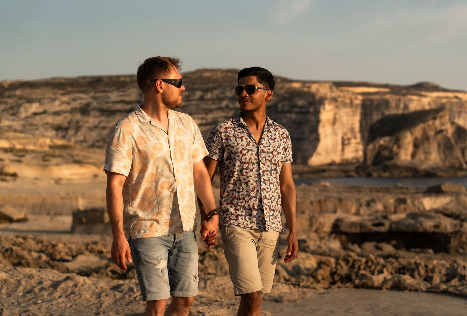 Two men holding hands and walking along a rocky coastal landscape at sunset wearing casual floral shirts and shorts.