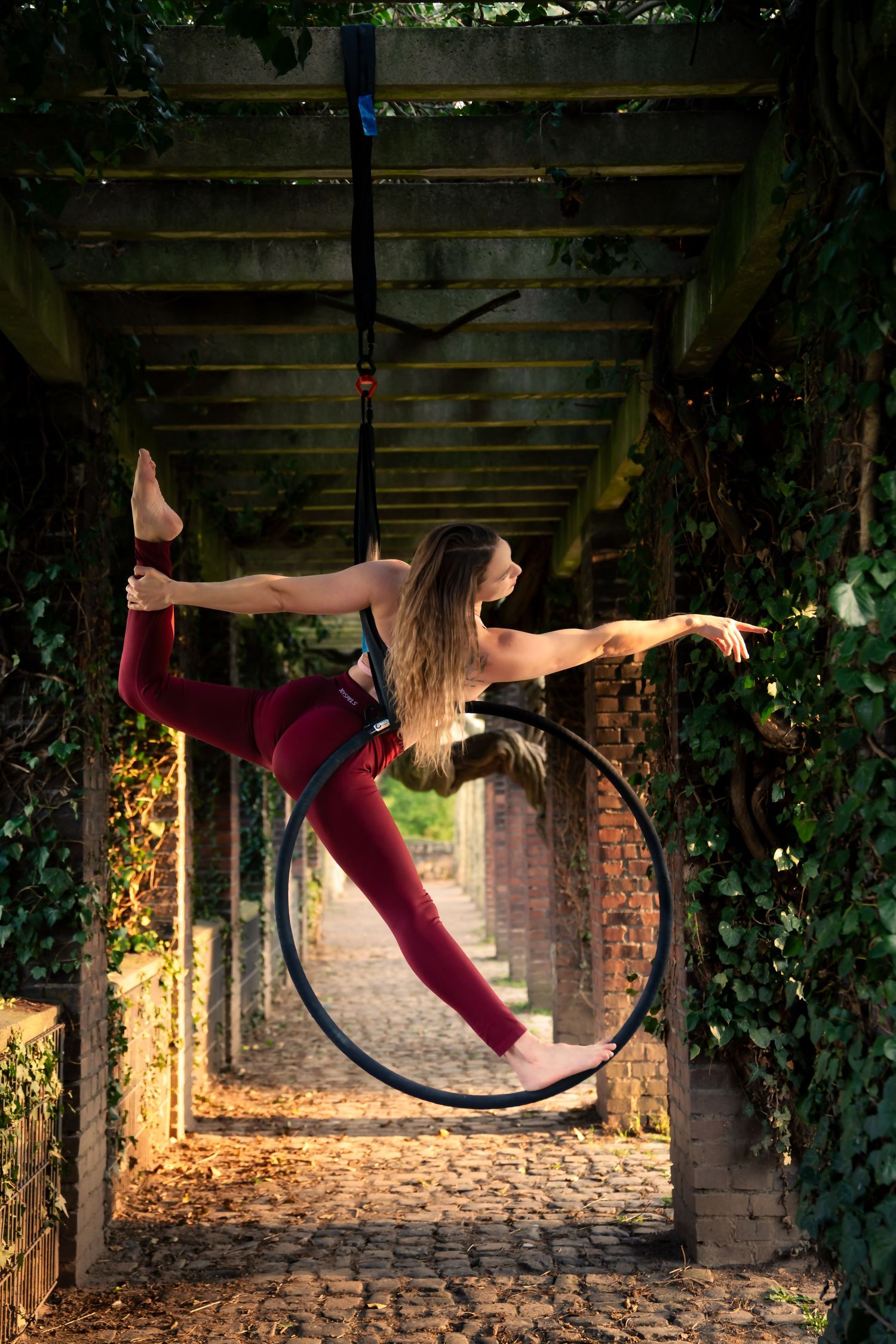 Woman in red performing aerial hoop acrobatics outdoors under a vine-covered pergola on a cobblestone path.