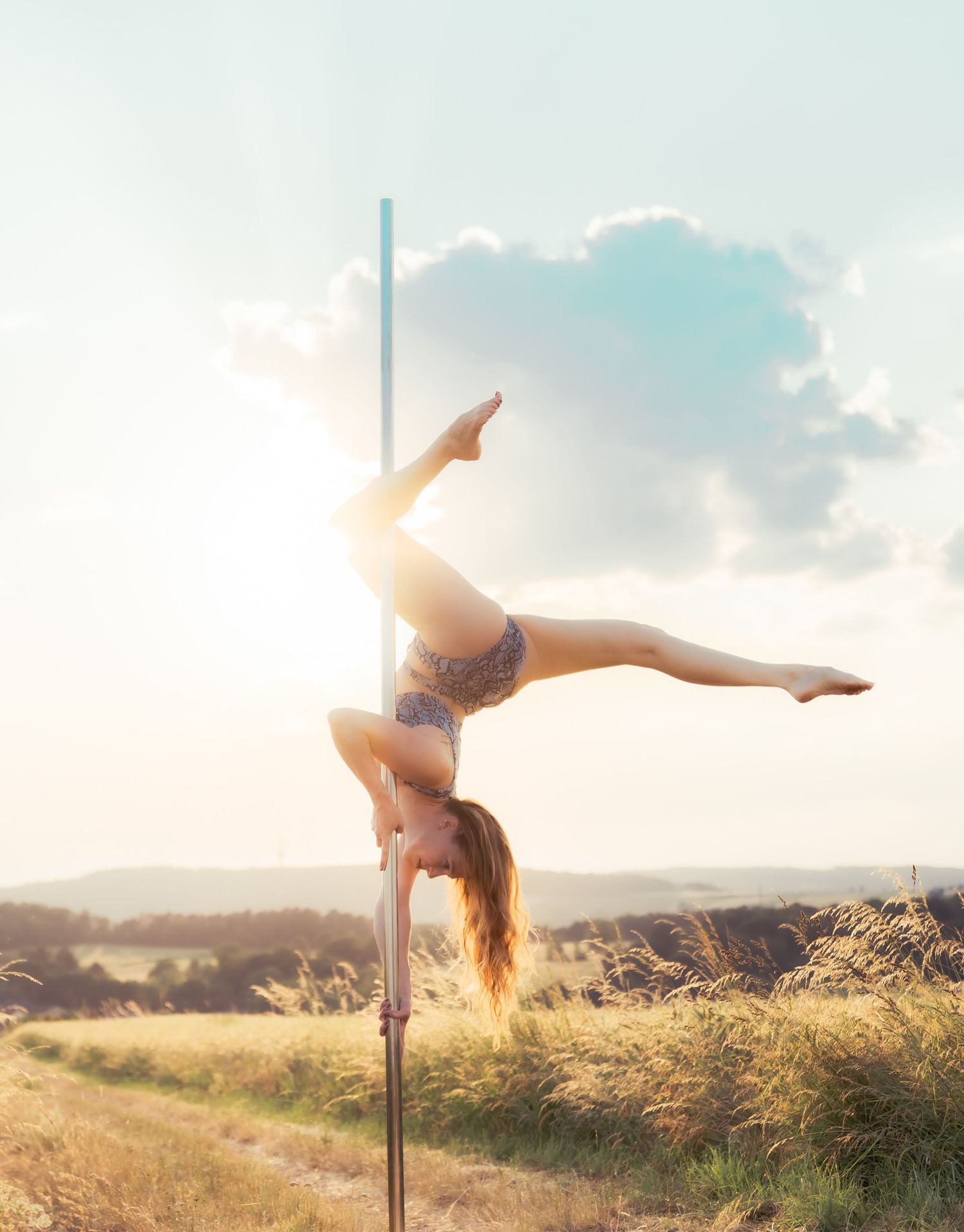 Woman performing an impressive pole dance split outdoors in a sunny field, showcasing strength and flexibility.