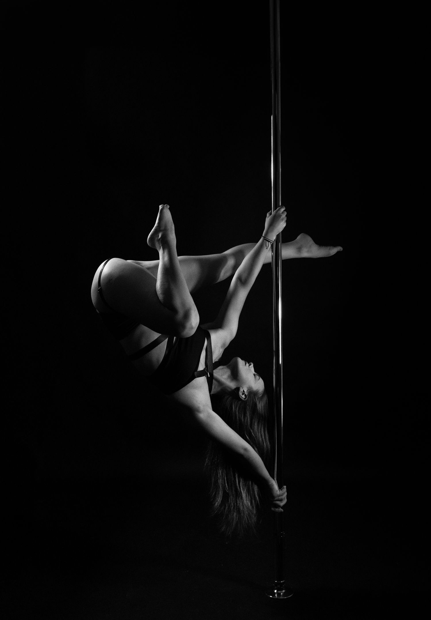 Black and white photo of a woman performing an inverted pole dance move, showcasing strength and flexibility.