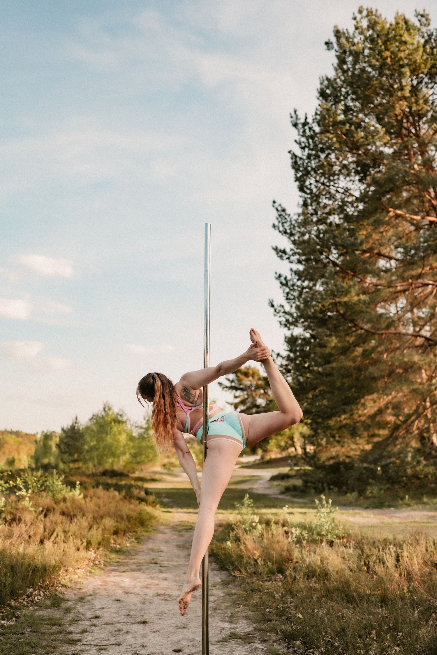 Flexible woman performing pole dance outdoors in nature, stretching leg high while holding pole in summer setting.