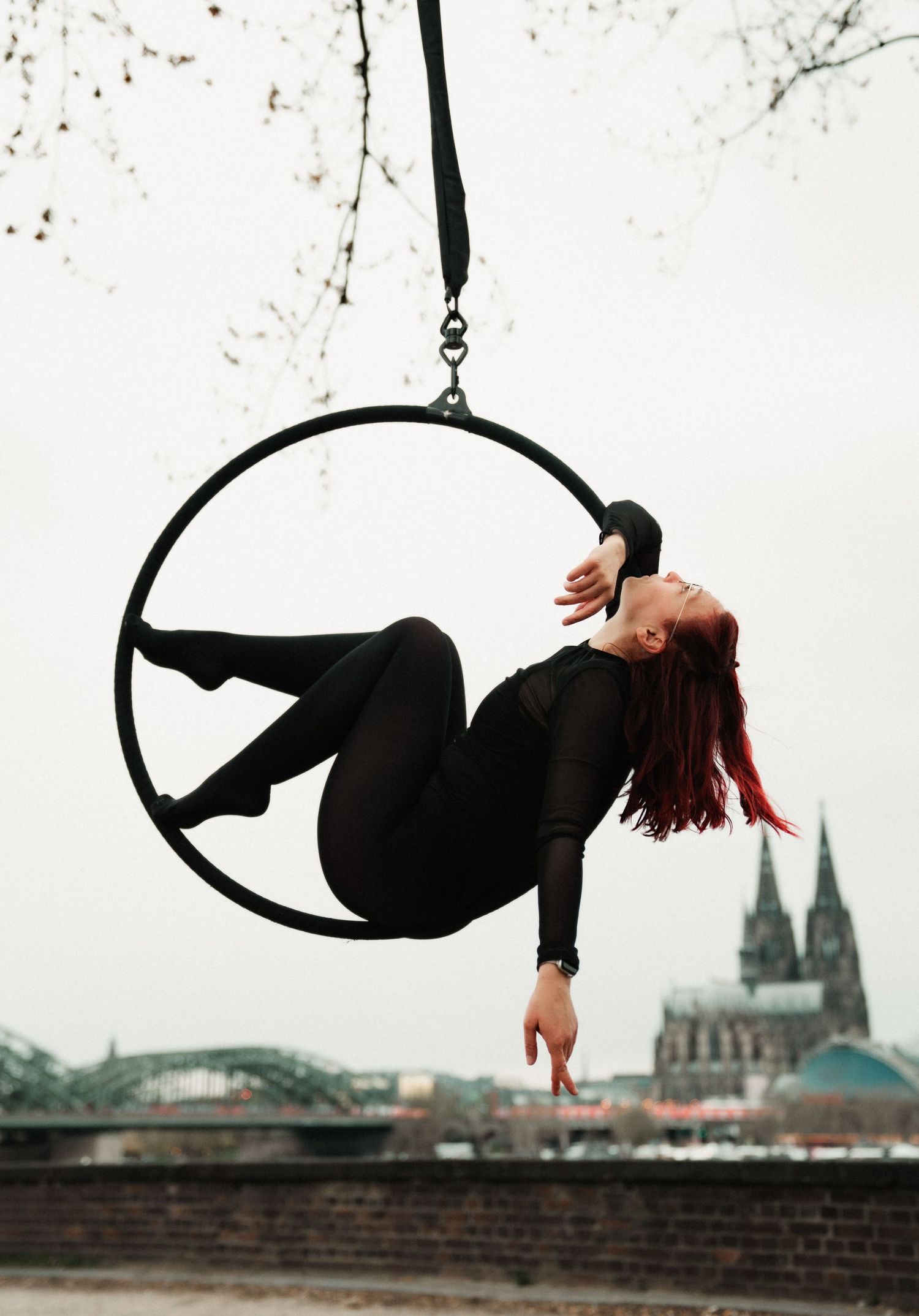 Aerial hoop performer in black outfit poses gracefully on lyra ring outdoors with Cologne Cathedral in background.