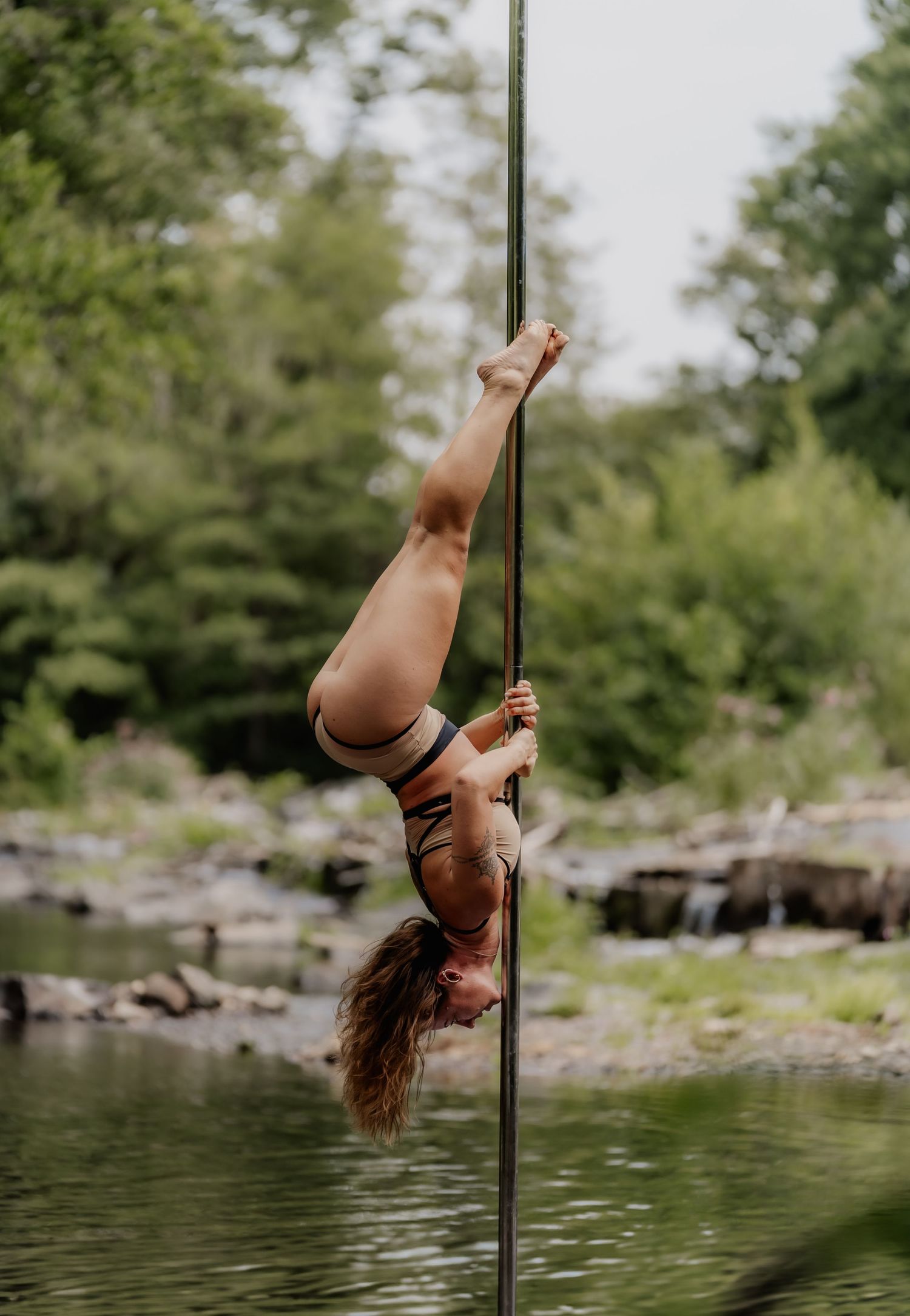 Athletic woman performing an inverted pole fitness move near a serene outdoor river setting.
