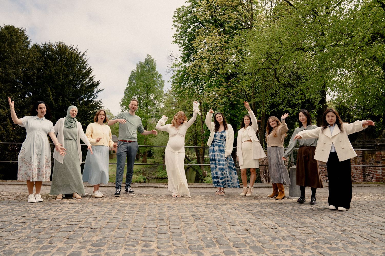 Group of ten people posing outdoors on cobblestone path with arms raised, surrounded by lush green trees.