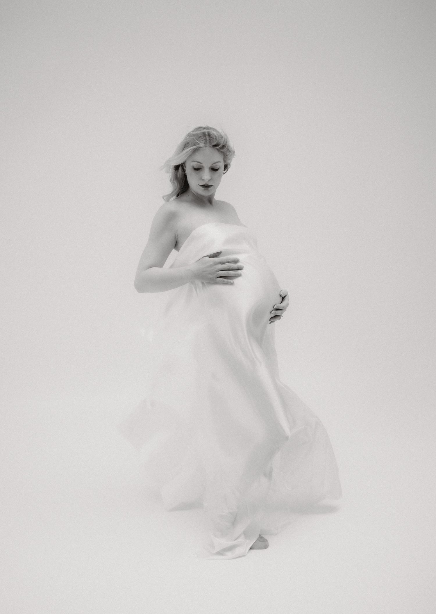 Black and white maternity photo of pregnant woman in flowing white fabric, cradling her baby bump in a studio setting.