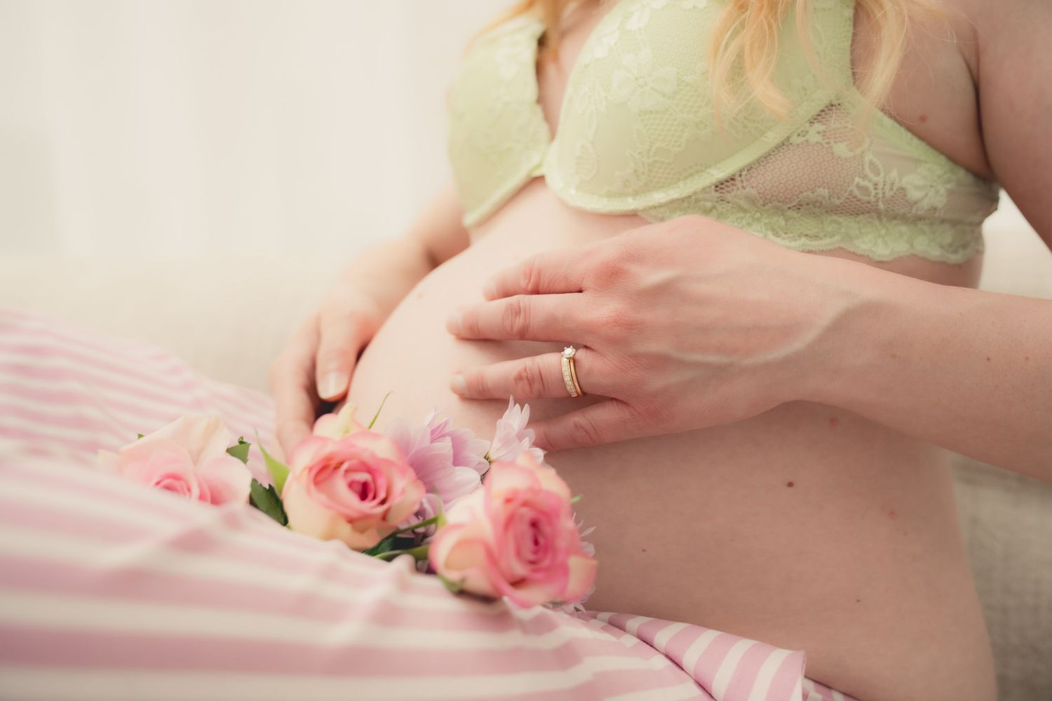 Pregnant woman in green lace bra cradling baby bump with pink roses on striped bedding.