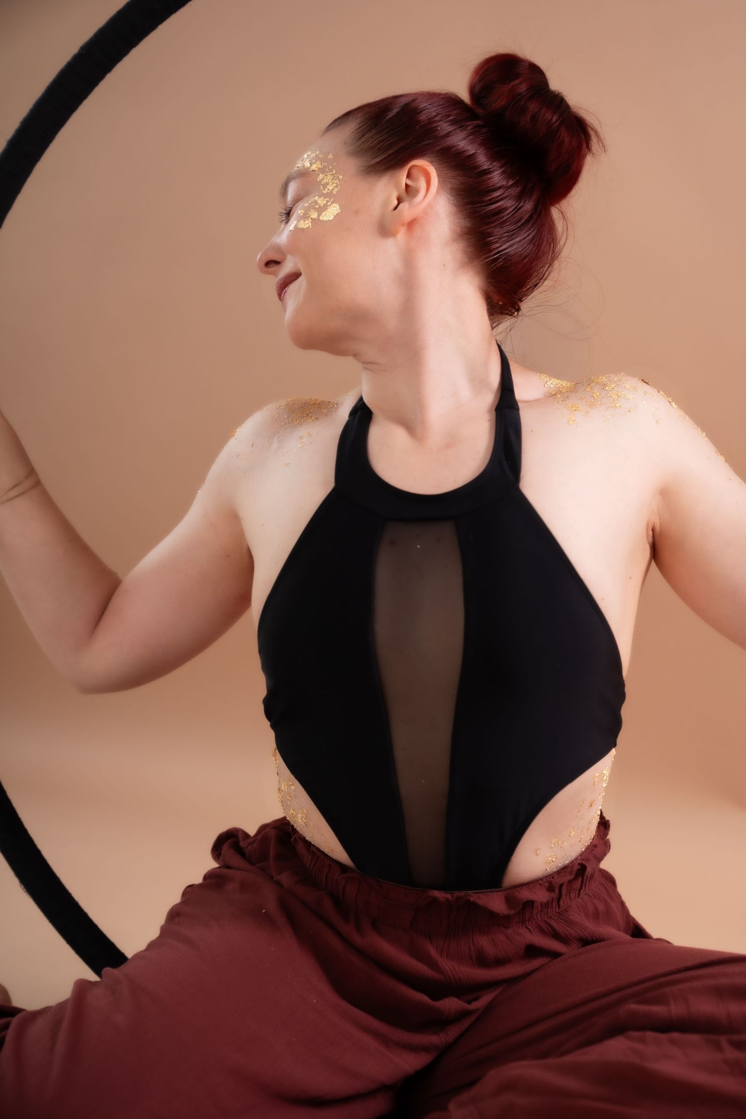 Woman with red hair and gold face paint poses gracefully in a black halter top and maroon pants inside a hoop.