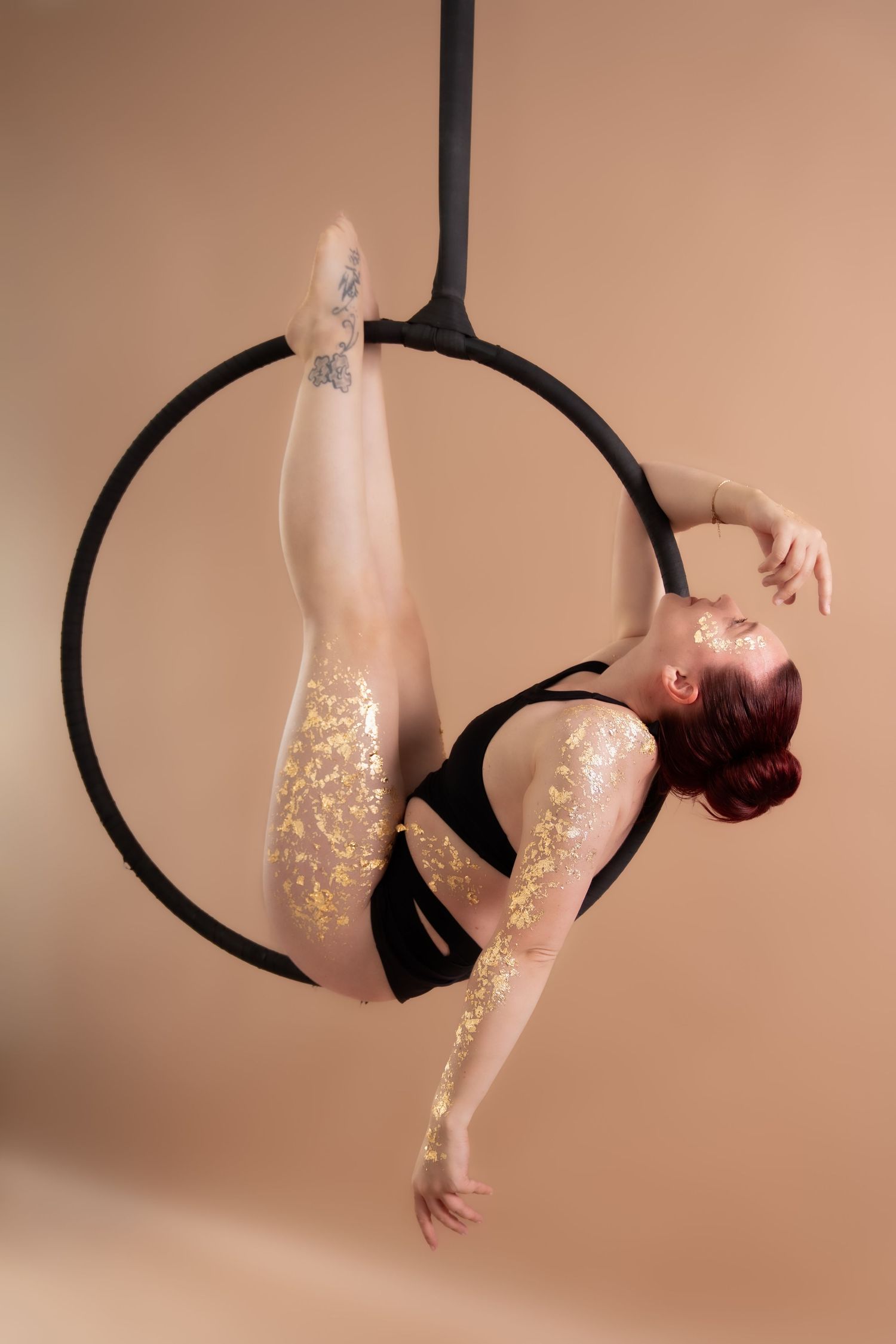A flexible aerial hoop performer in black leotard with gold glitter poses gracefully on a lyra ring against a beige background.