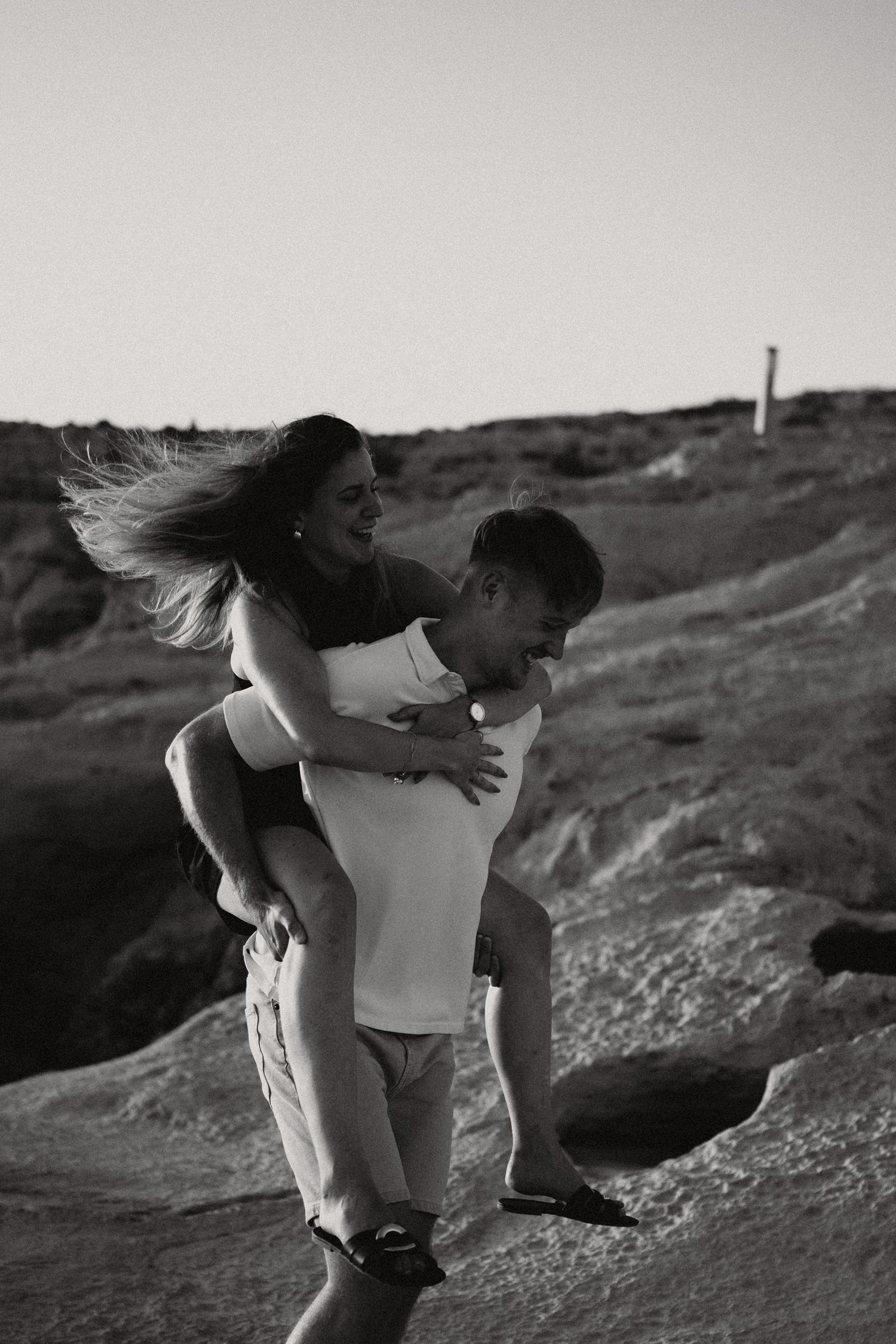 Black and white photo of a man giving a woman a piggyback ride on rocky terrain, her hair blowing in the wind.
