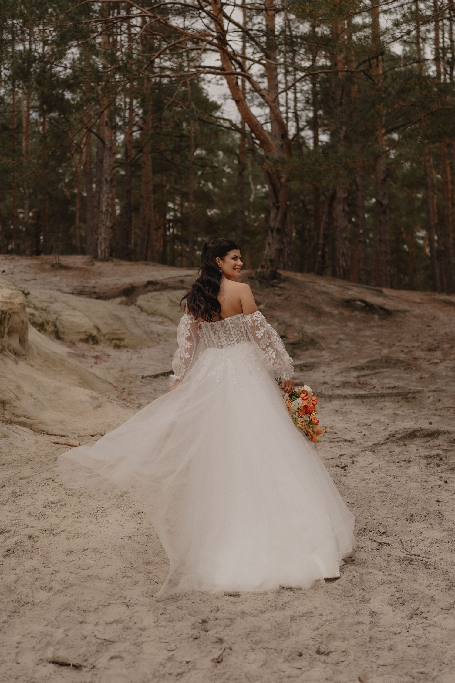 A flowing white wedding dress twirls in a pine forest setting with sandy ground.