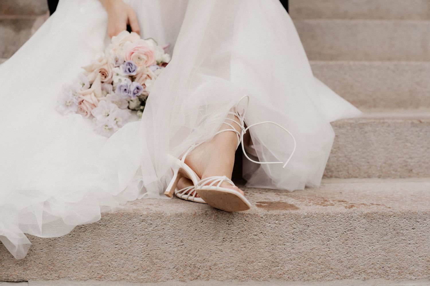 Elegant bridal shoes and flowing white wedding dress captured on stone steps during a romantic outdoor photoshoot.
