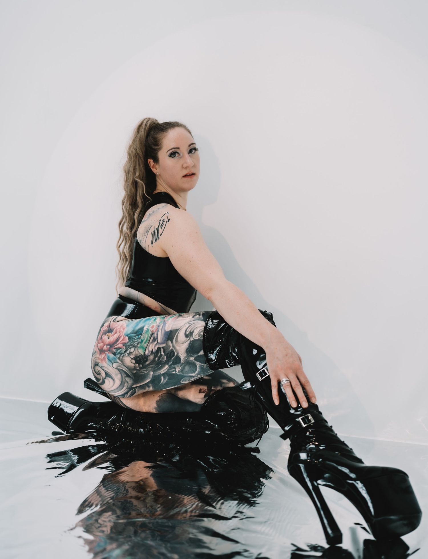 A person in black clothing and platform boots poses against a white wall with a floral print bag.