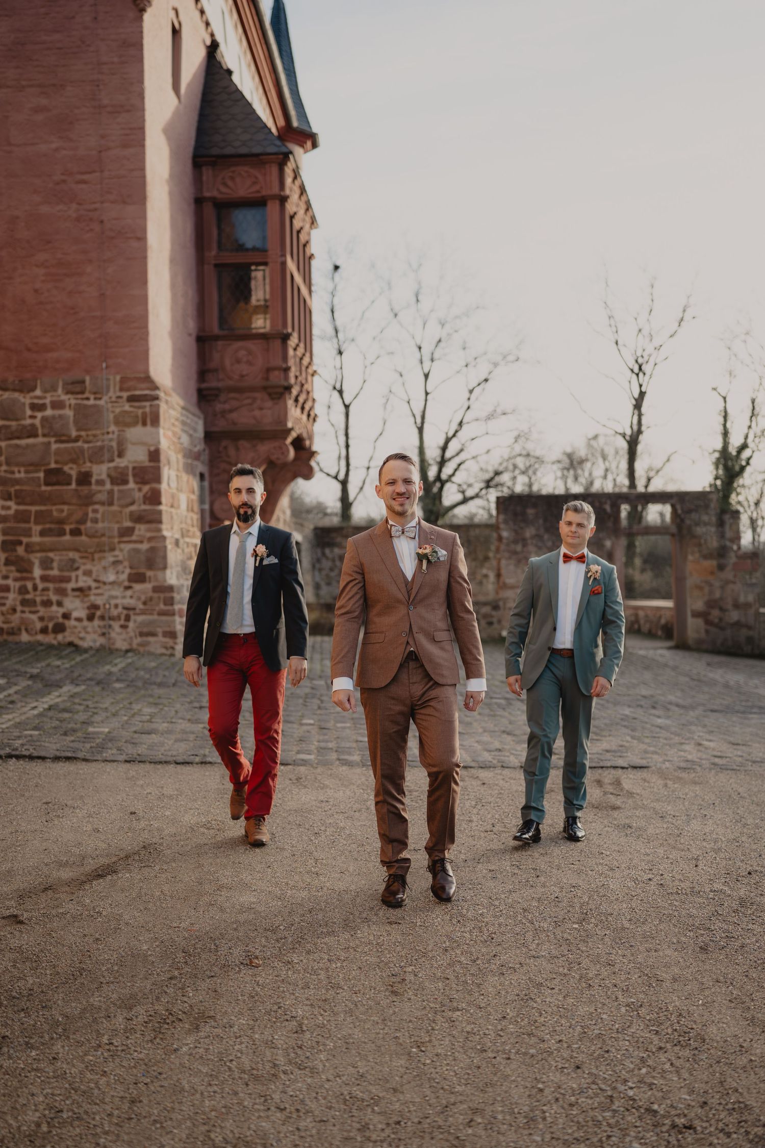 Groomsmen in brown and blue suits walk together outside a historic venue.