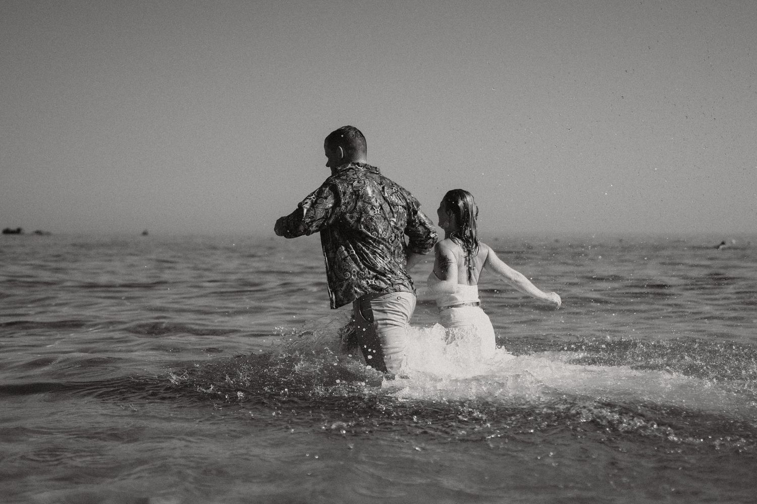 Black and white image shows couple splashing through ocean waves during seaside shoot.