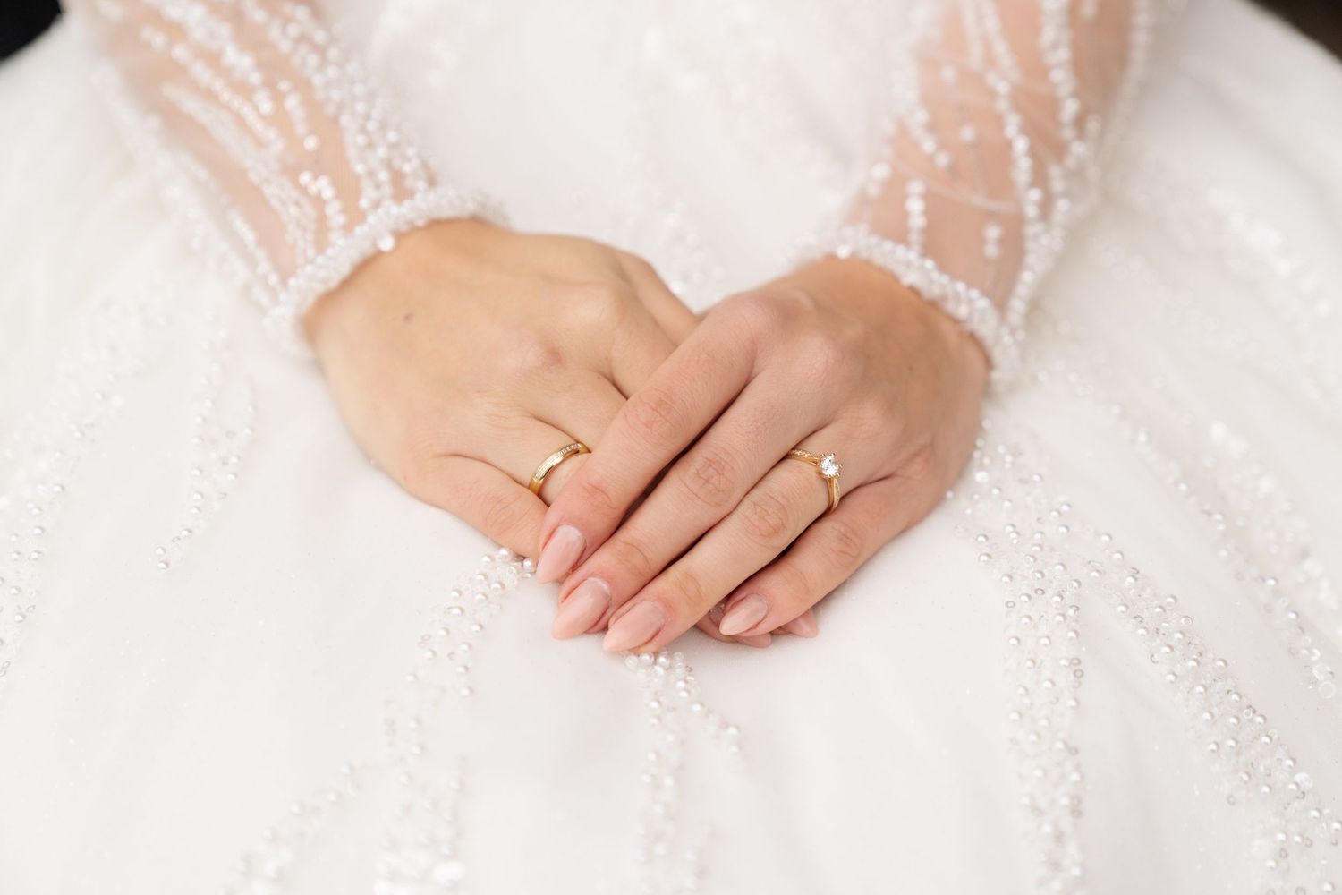 Delicate details of lace and beading on a white wedding dress with hands gently resting on fabric.