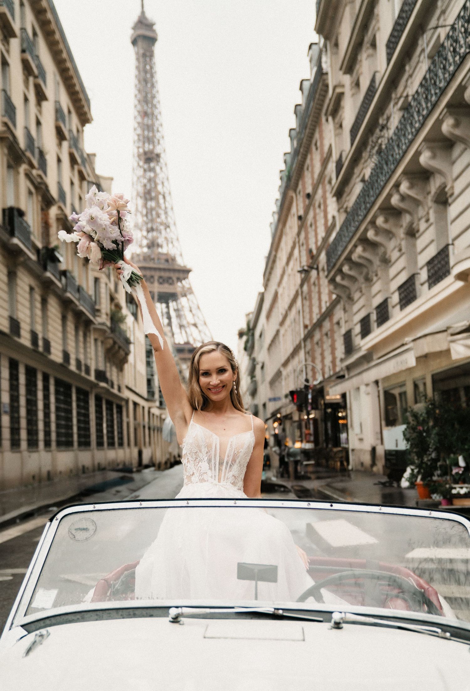 A bride in a white dress poses in a vintage car on a charming Parisian street with the Eiffel Tower visible in the background.