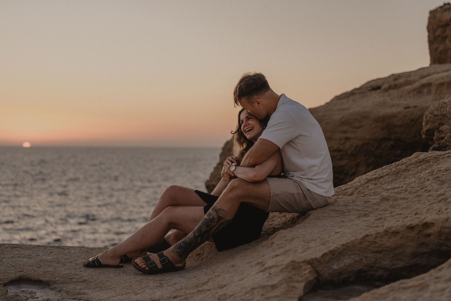 Couple sitting together on rocky cliffside at sunset by the ocean, sharing a romantic embrace.