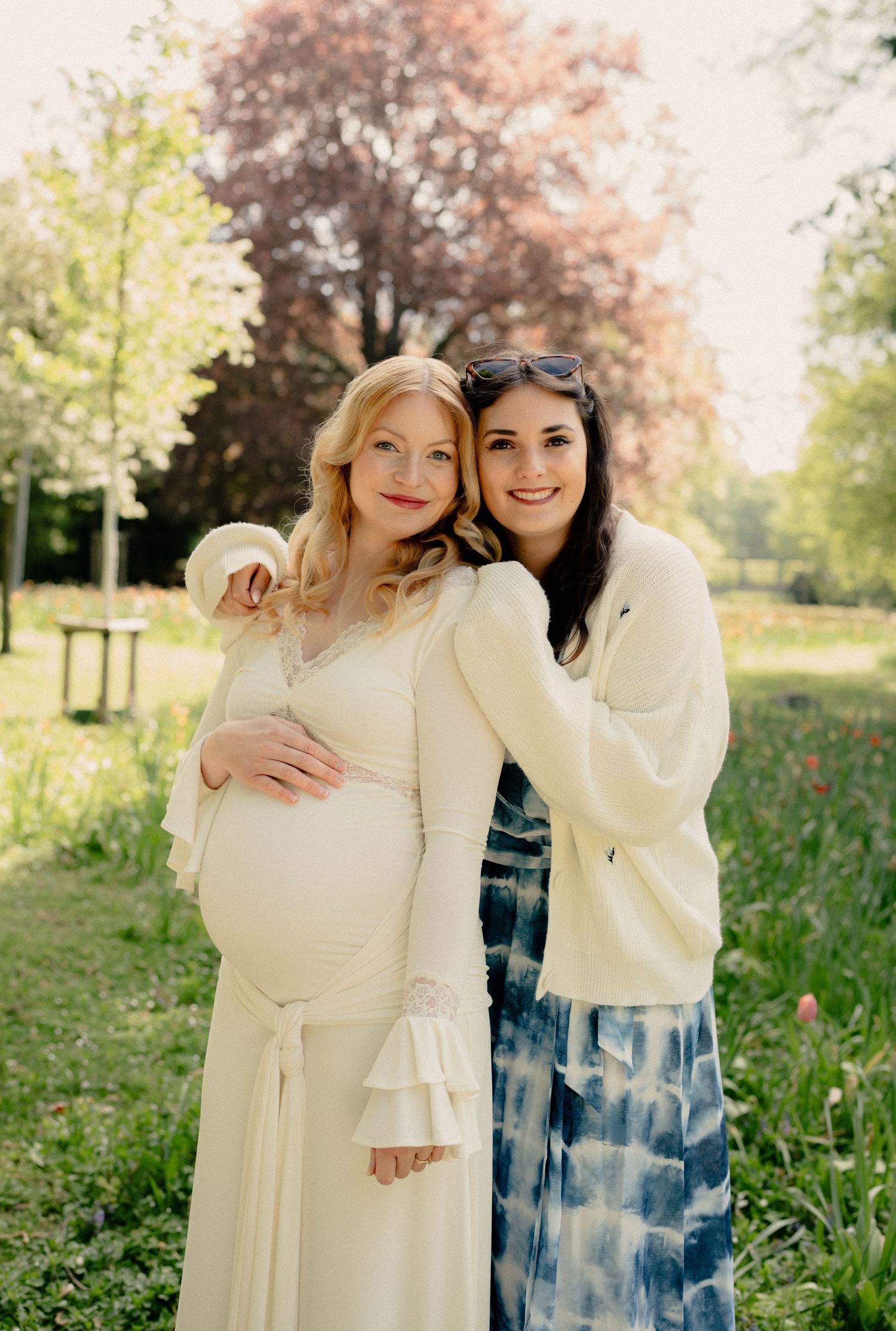 Pregnant woman in white dress posing with a dark-haired friend in a sunny green park.