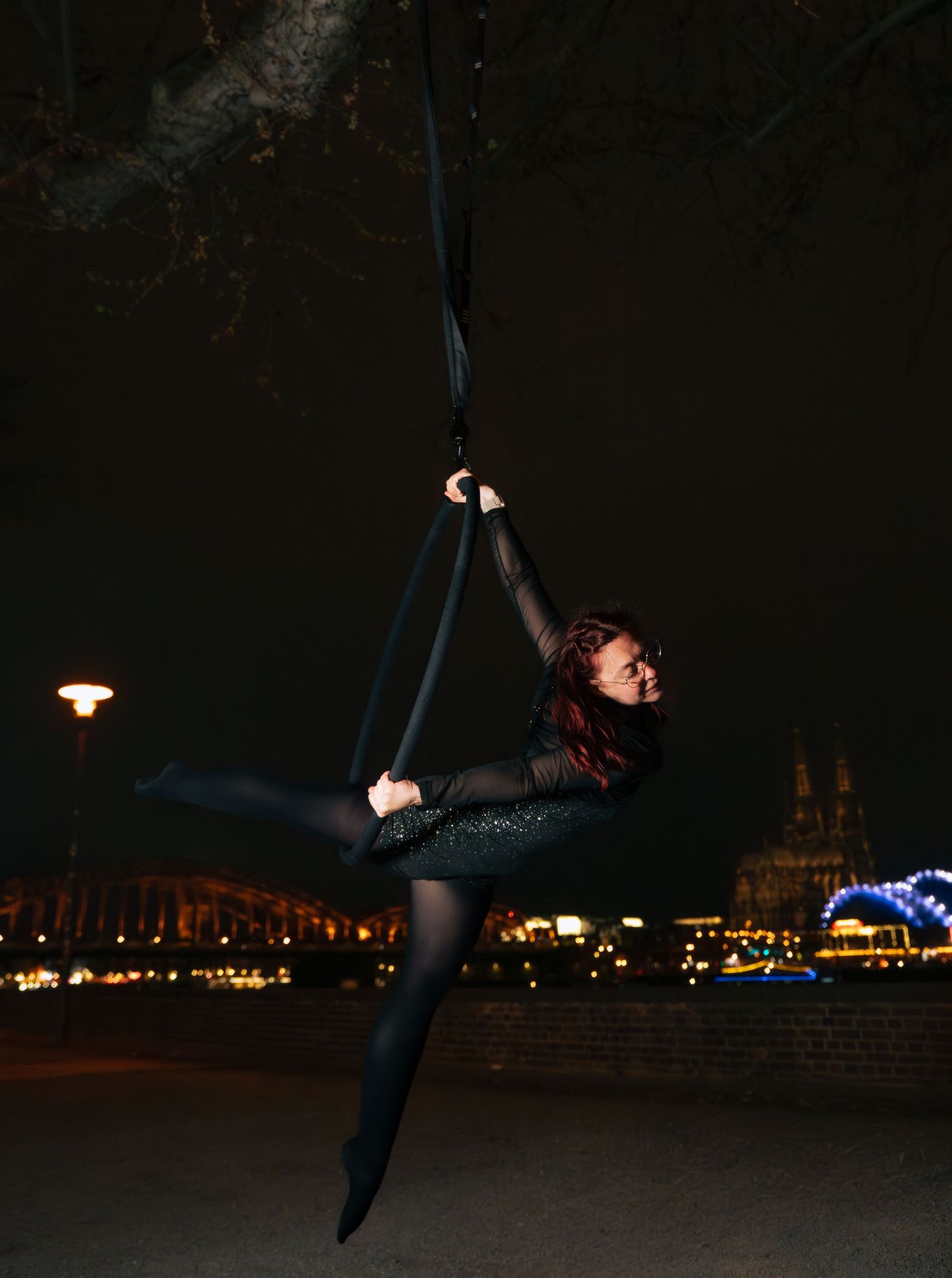Aerial silk performer in black outfit gracefully poses mid-air at night with city lights and cathedral in background.