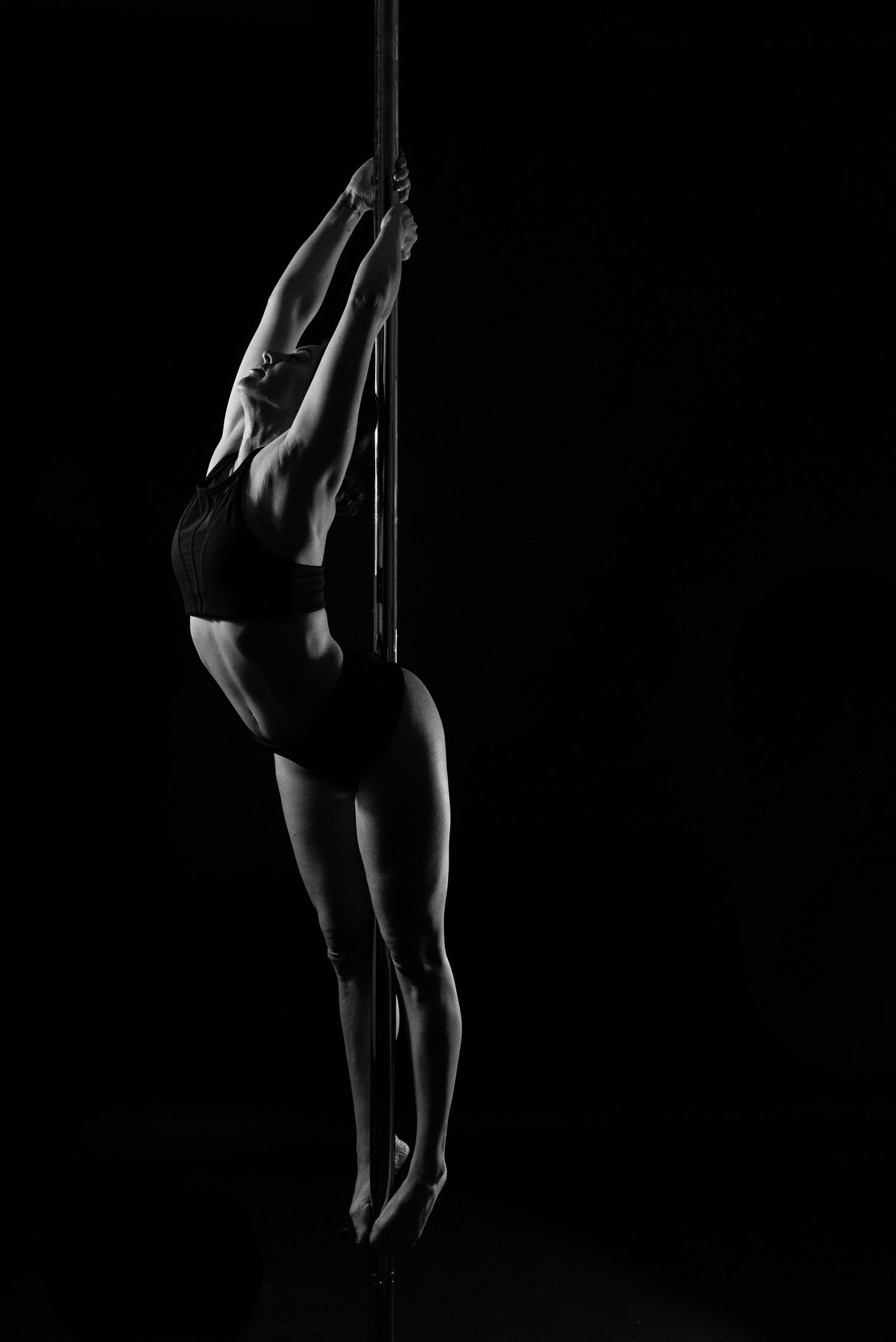 Black and white photo of a female pole dancer gracefully suspended on a pole against a dark background.