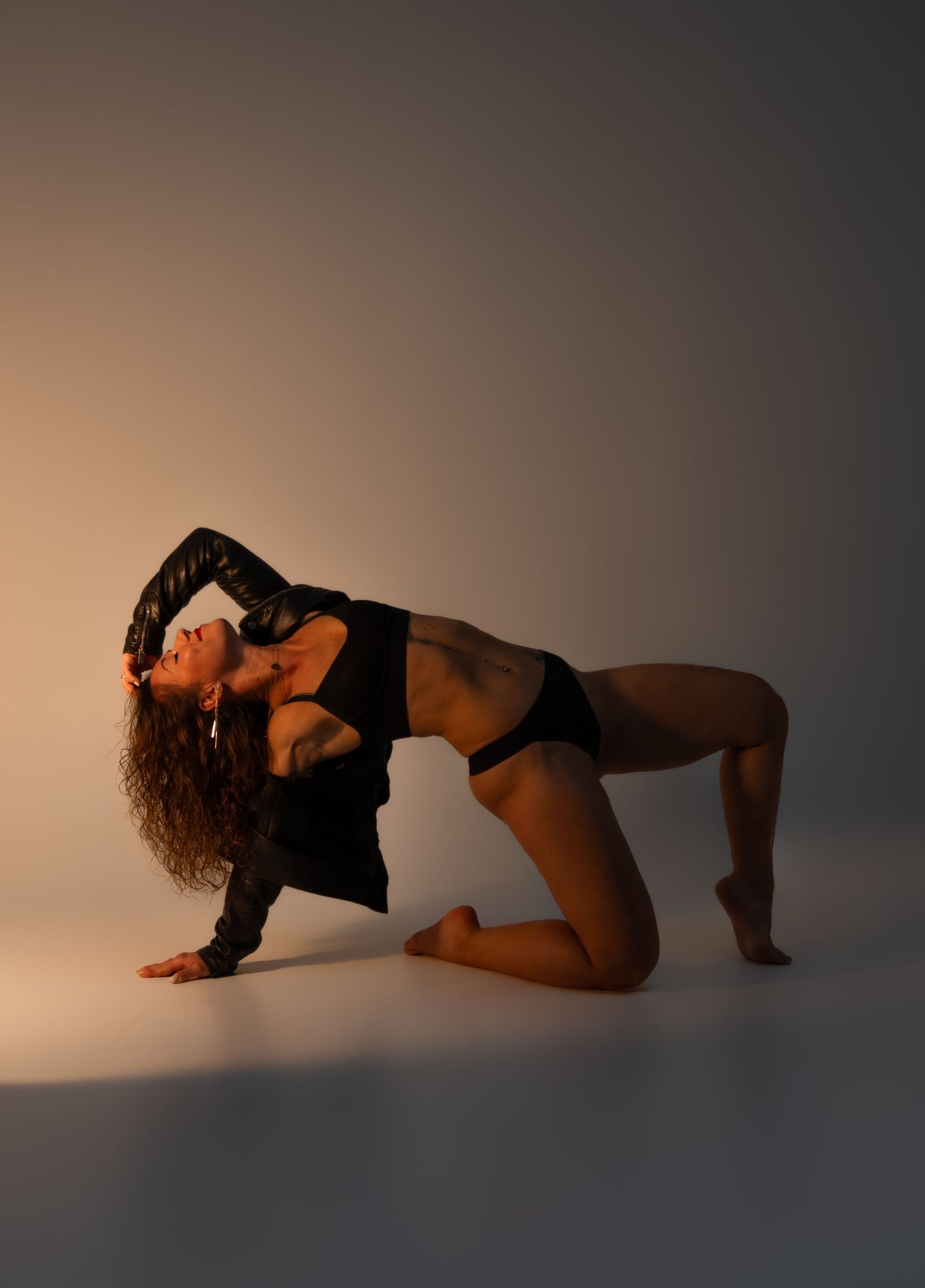 A female dancer in black attire poses on knees arching back gracefully in a dramatic studio dance pose.