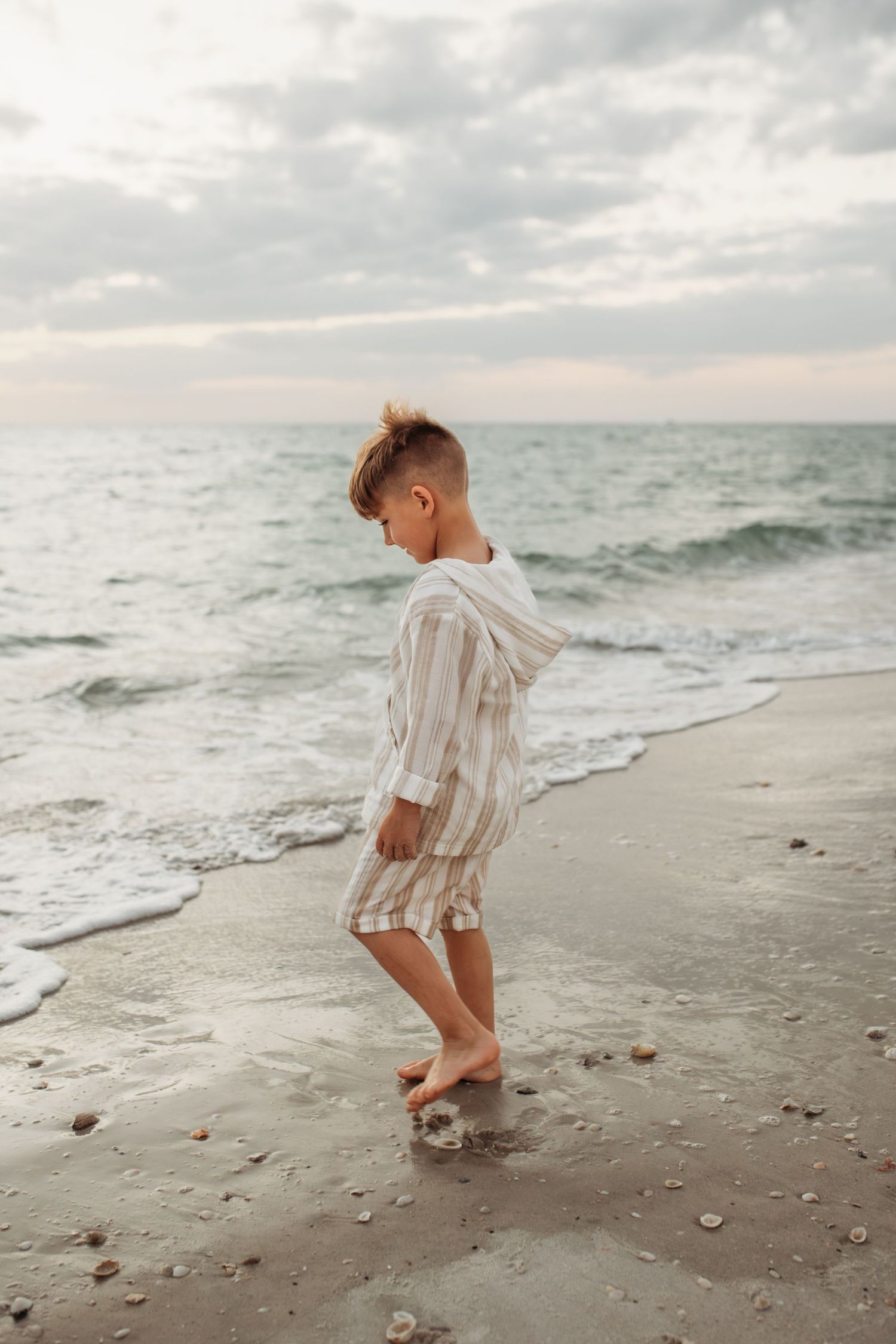 Young boy in striped linen outfit walking barefoot on sandy beach shore with gentle ocean waves at sunset.
