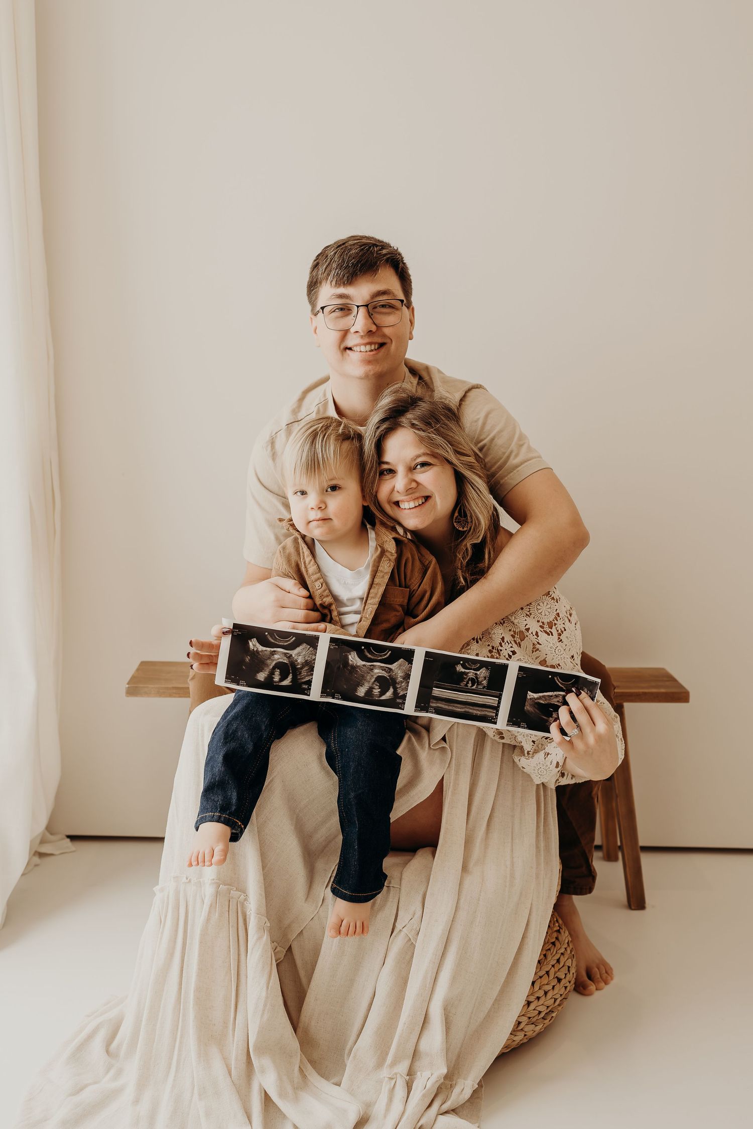 Happy family of three holding ultrasound photos on a bench, announcing a new pregnancy in a bright studio setting.
