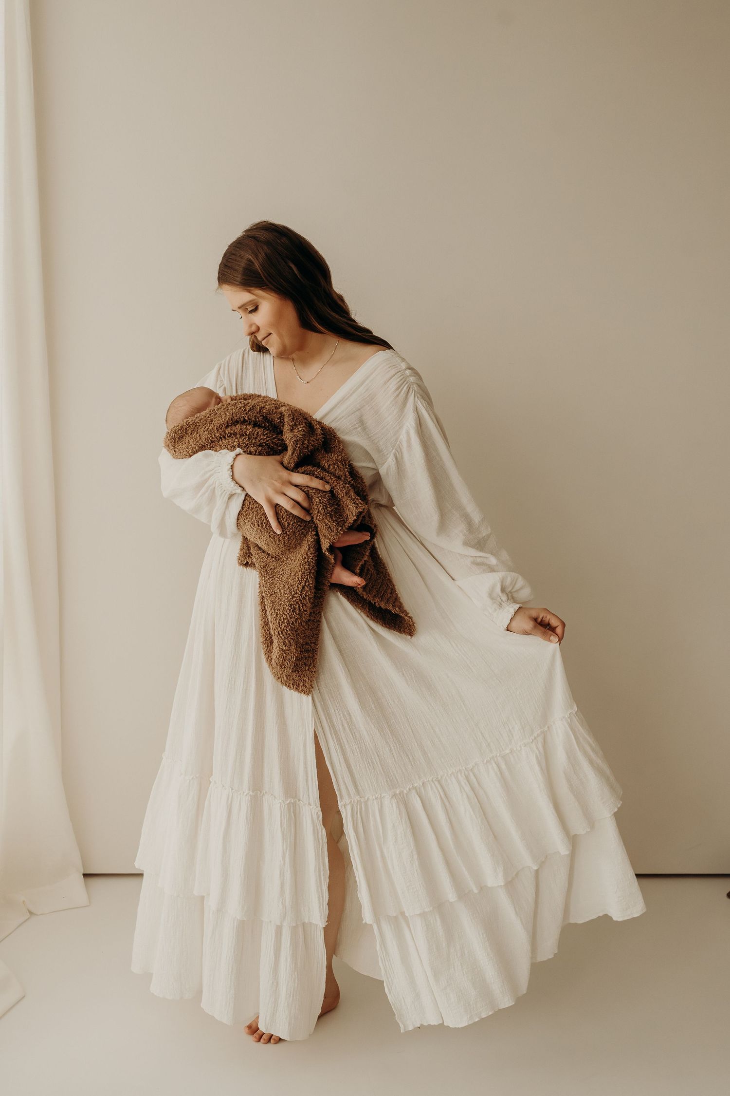 A mother in a white tiered dress tenderly holds her newborn wrapped in a brown fuzzy blanket in a bright studio.