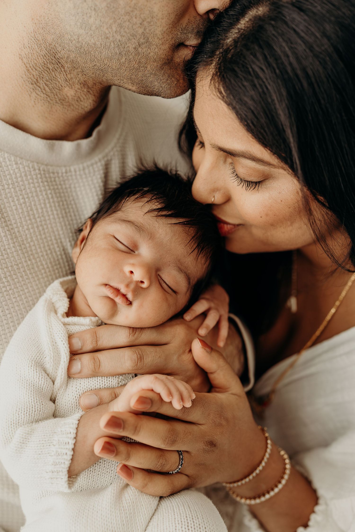 Parents kissing their sleeping newborn baby wrapped in white blanket, tender family portrait.
