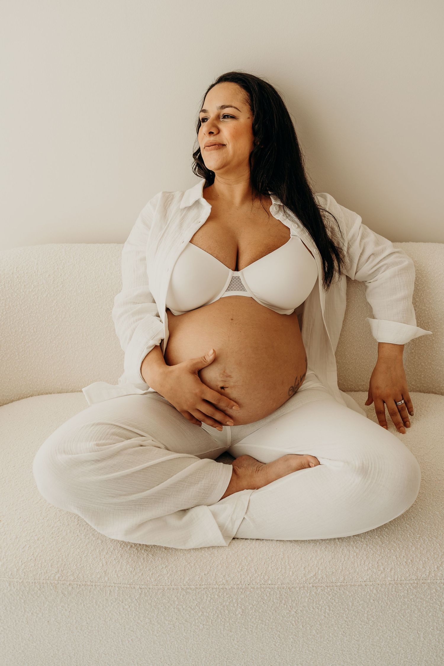 A minimalist maternity photoshoot featuring a woman in white clothing reclining on a light-colored backdrop.