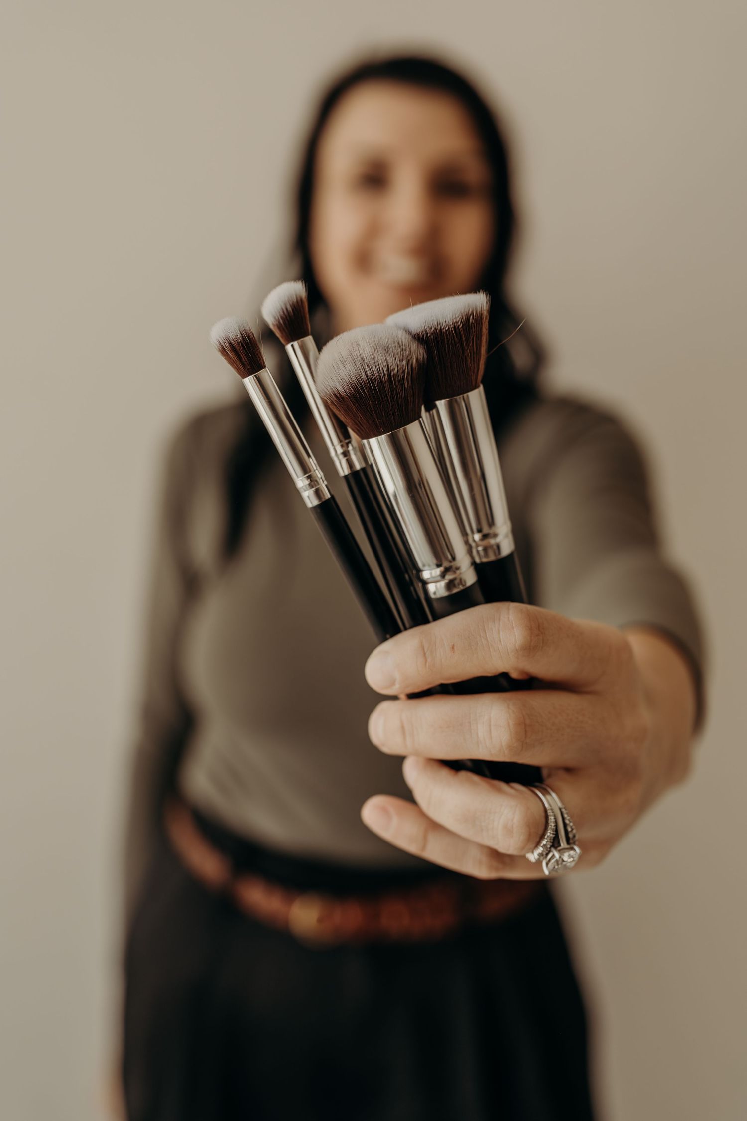 A hand holds up a collection of makeup brushes against a neutral background in a minimalist style.
