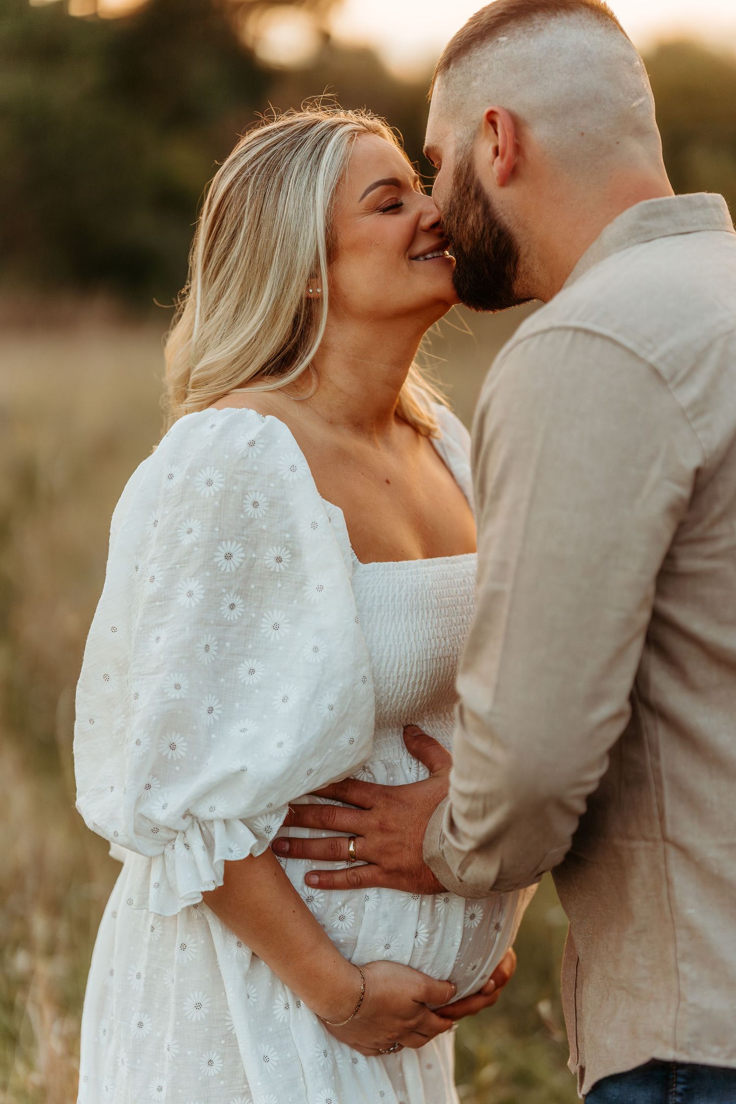 Couple in white and beige sharing romantic moment in golden field at sunset during their maternity session.