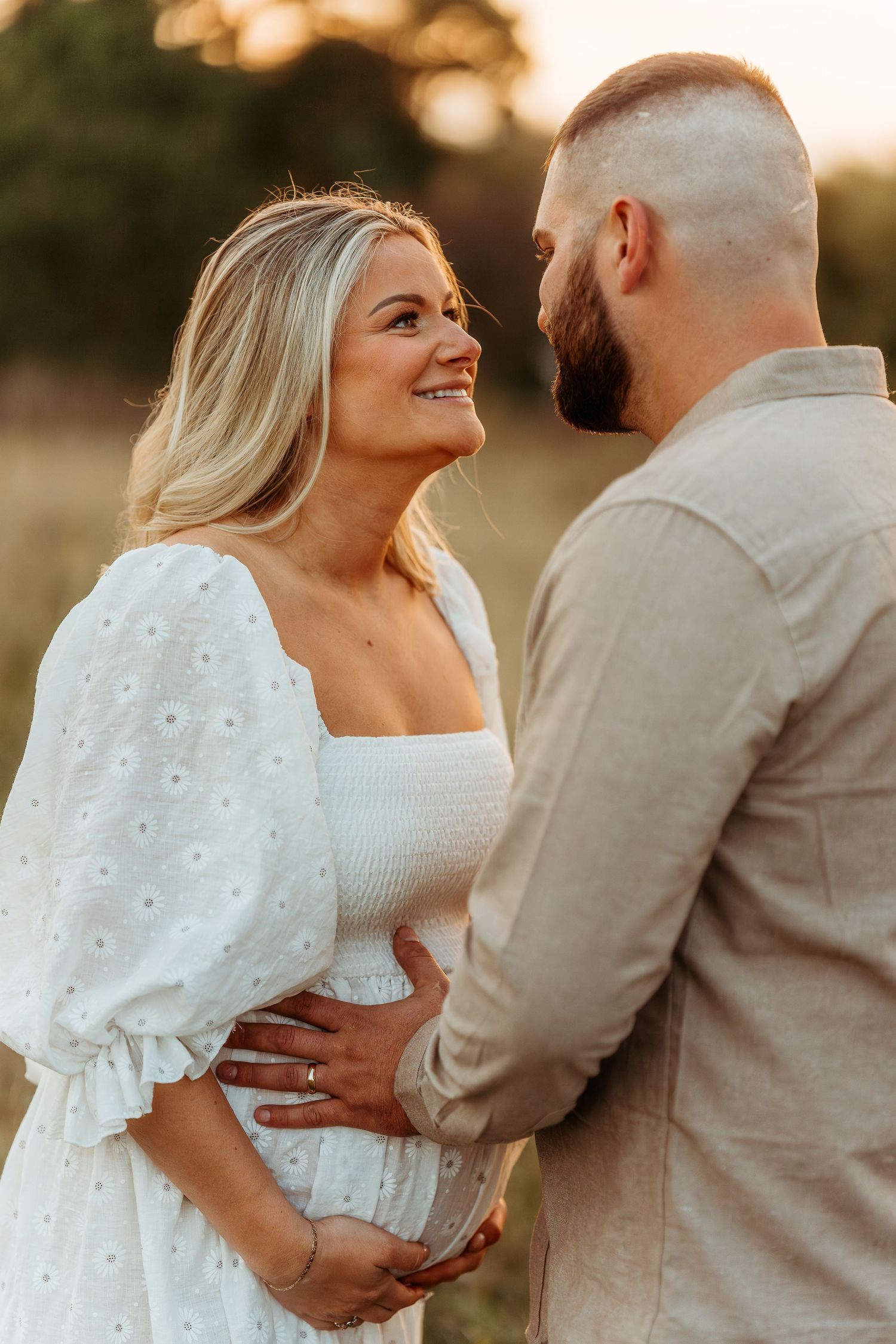 Romantic couple embracing in a field at golden hour wearing white and beige outfits showcasing pregnancy.