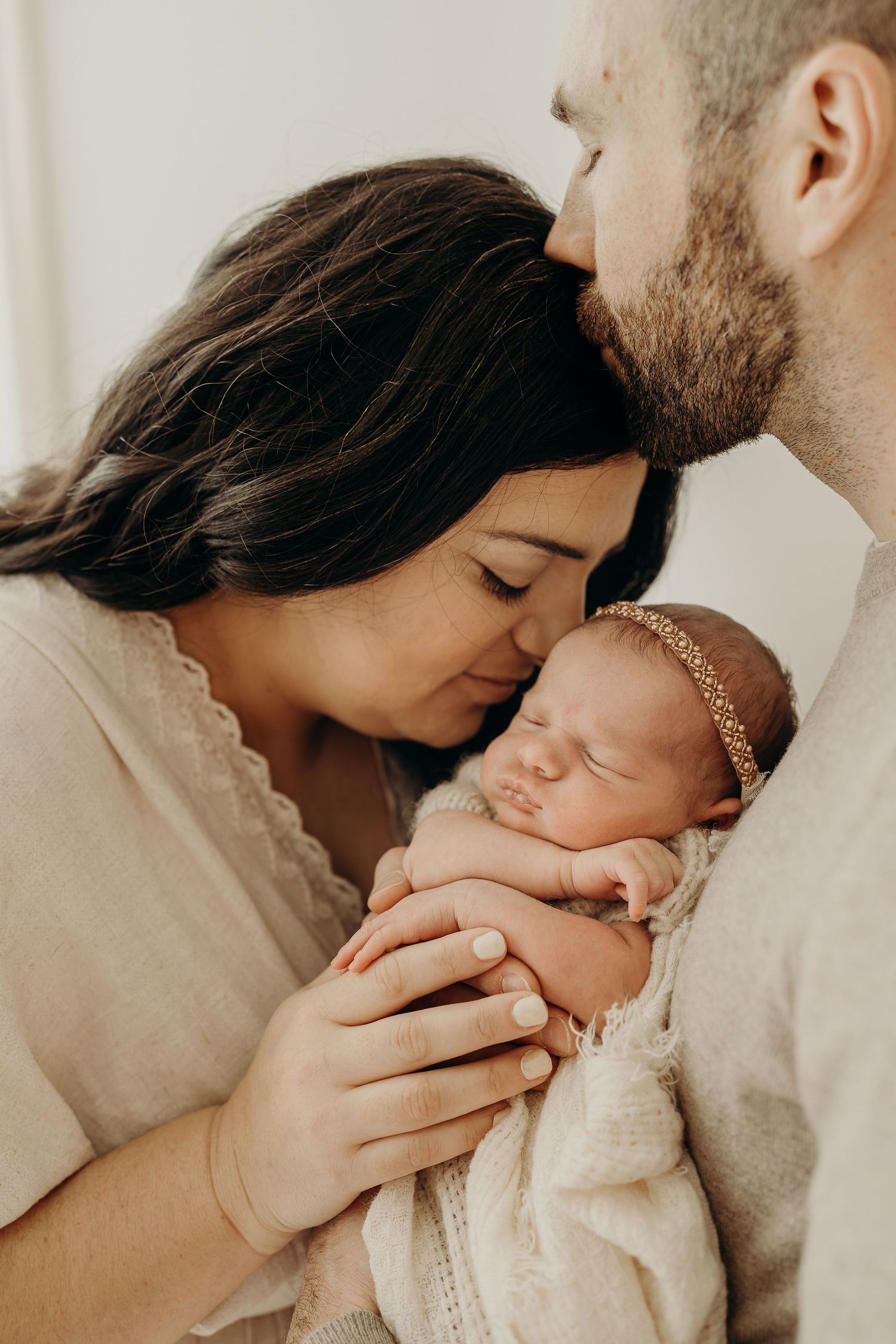 New parents tenderly kiss their sleeping newborn baby wrapped in a cream swaddle with a gold headband at Slatestudio in Wisconsin