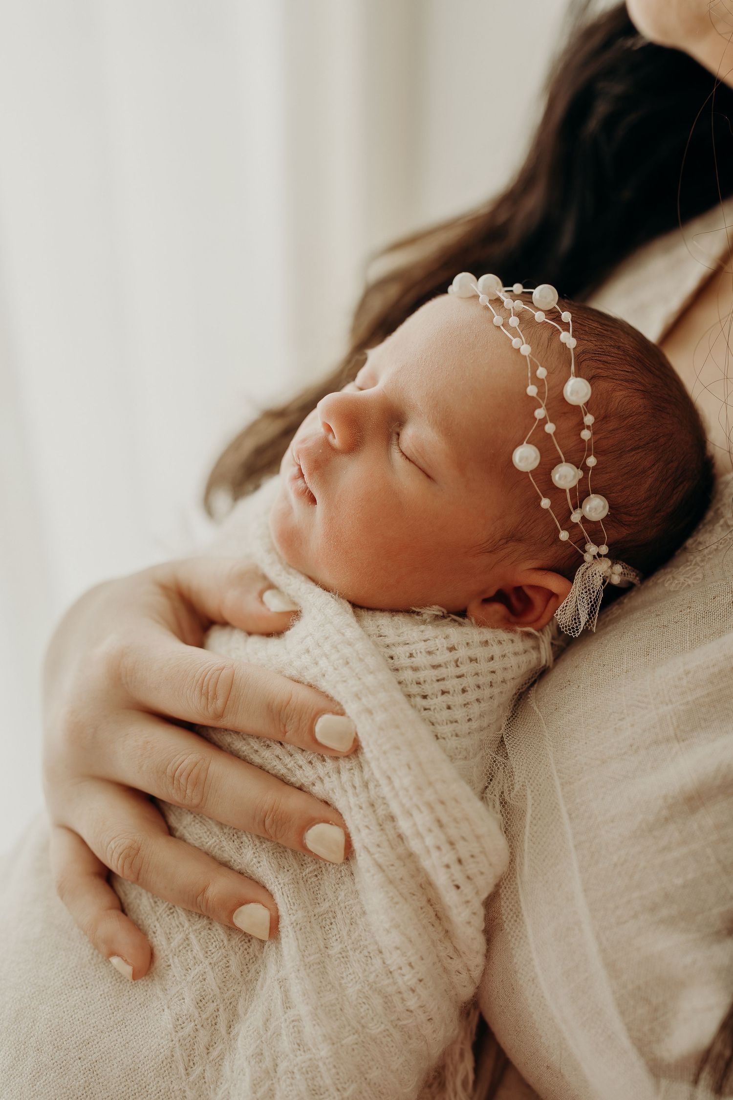Newborn baby sleeping peacefully in mother's arms, wrapped in white knit blanket with delicate pearl headband.