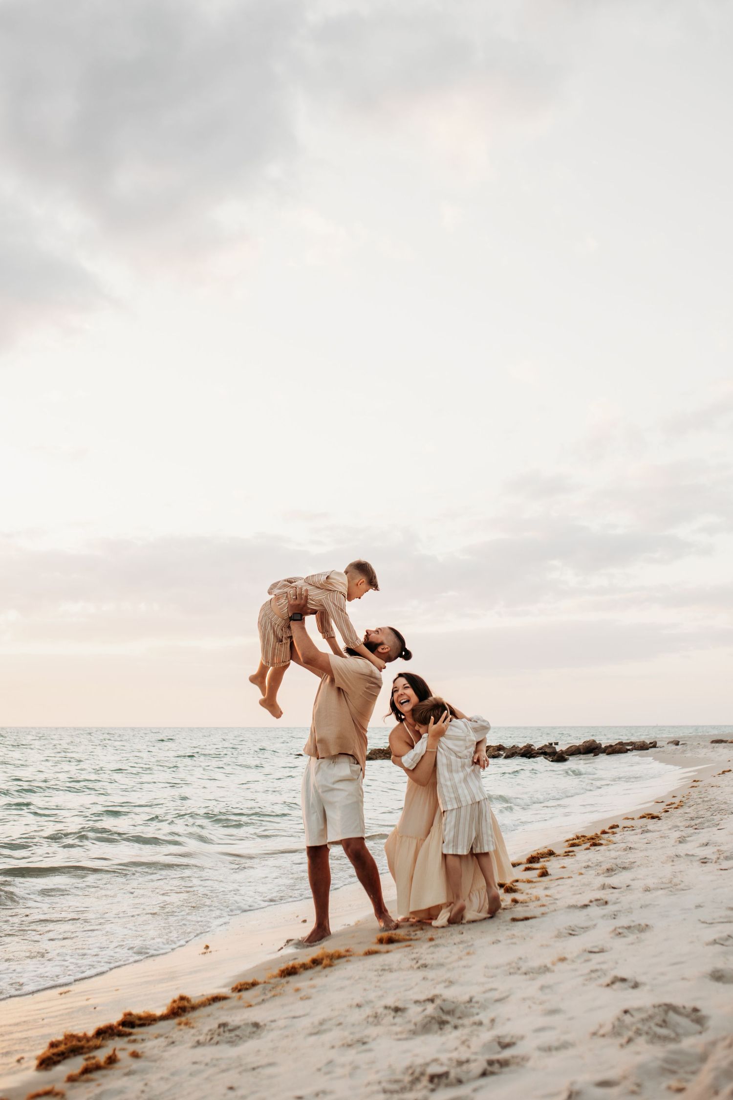 Family enjoying a beach sunset, father lifting child in air while mother holds baby on sandy shore.