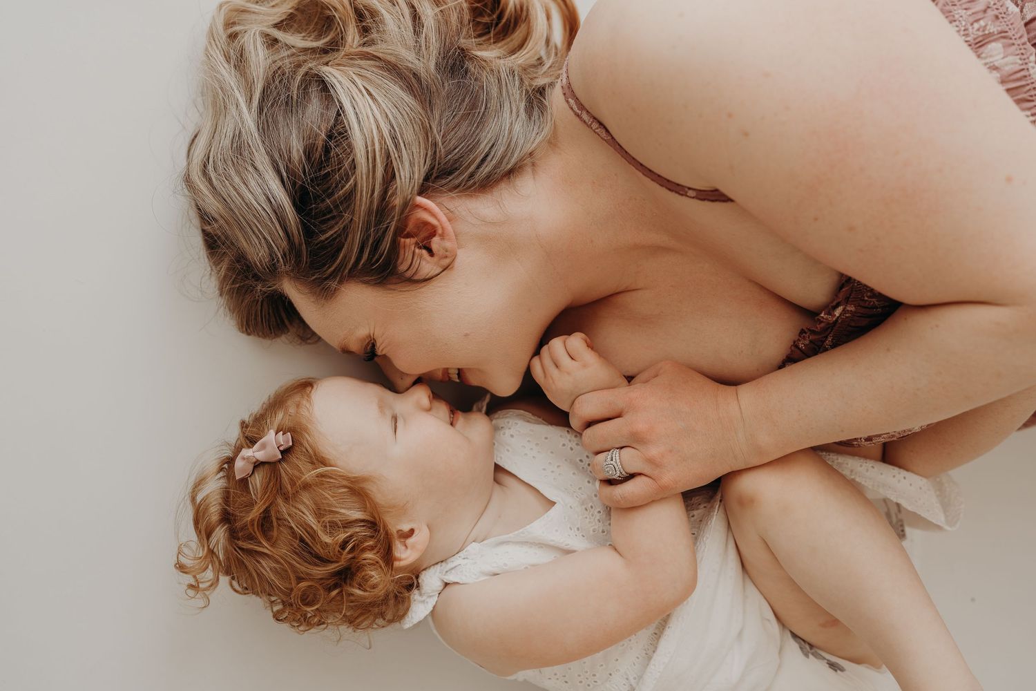 A mother tenderly nuzzles her curly-haired baby girl while lying together on a soft light surface.