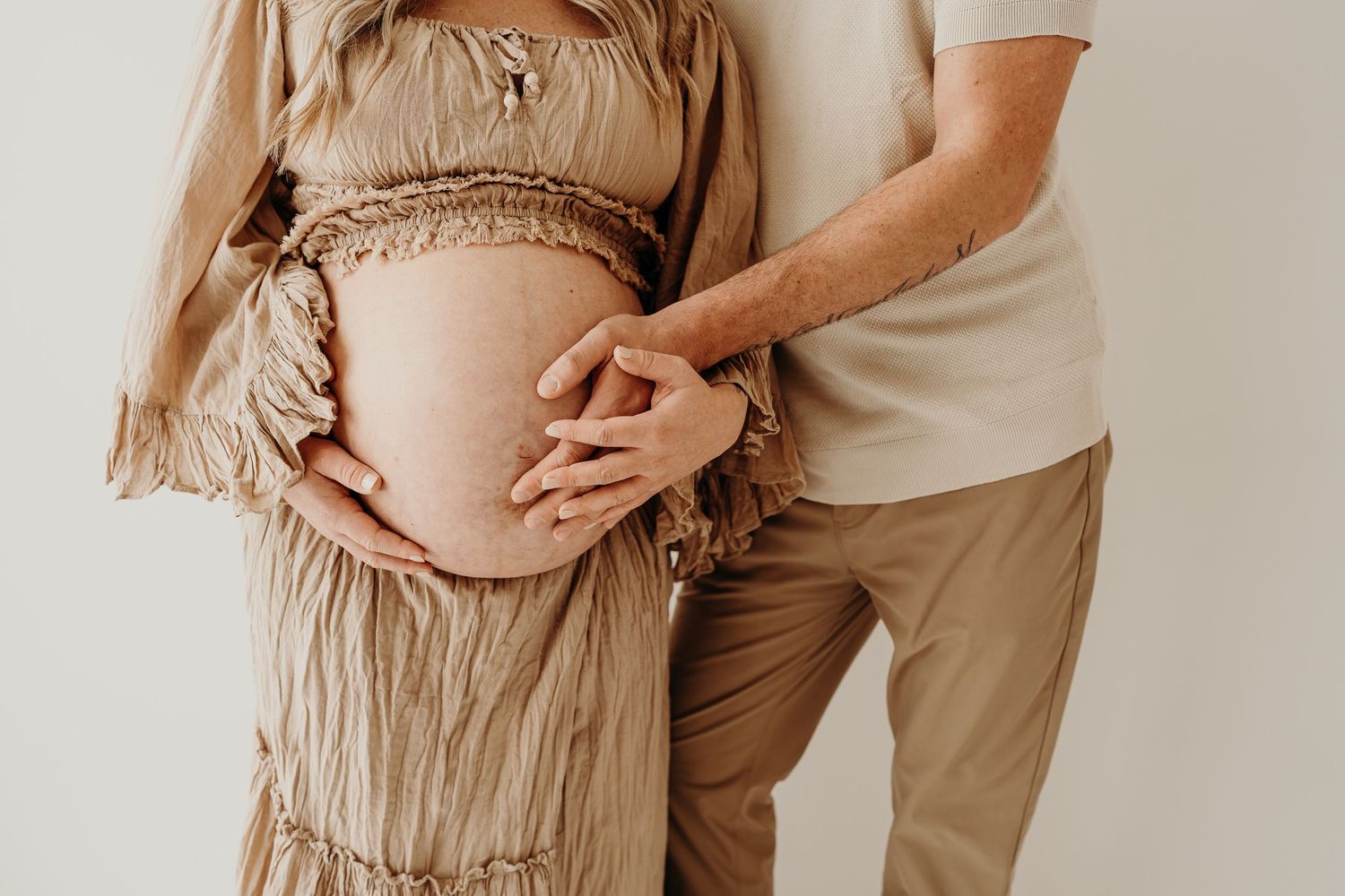 mom and dad standing by a studio window with hands on mom's pregnant belly