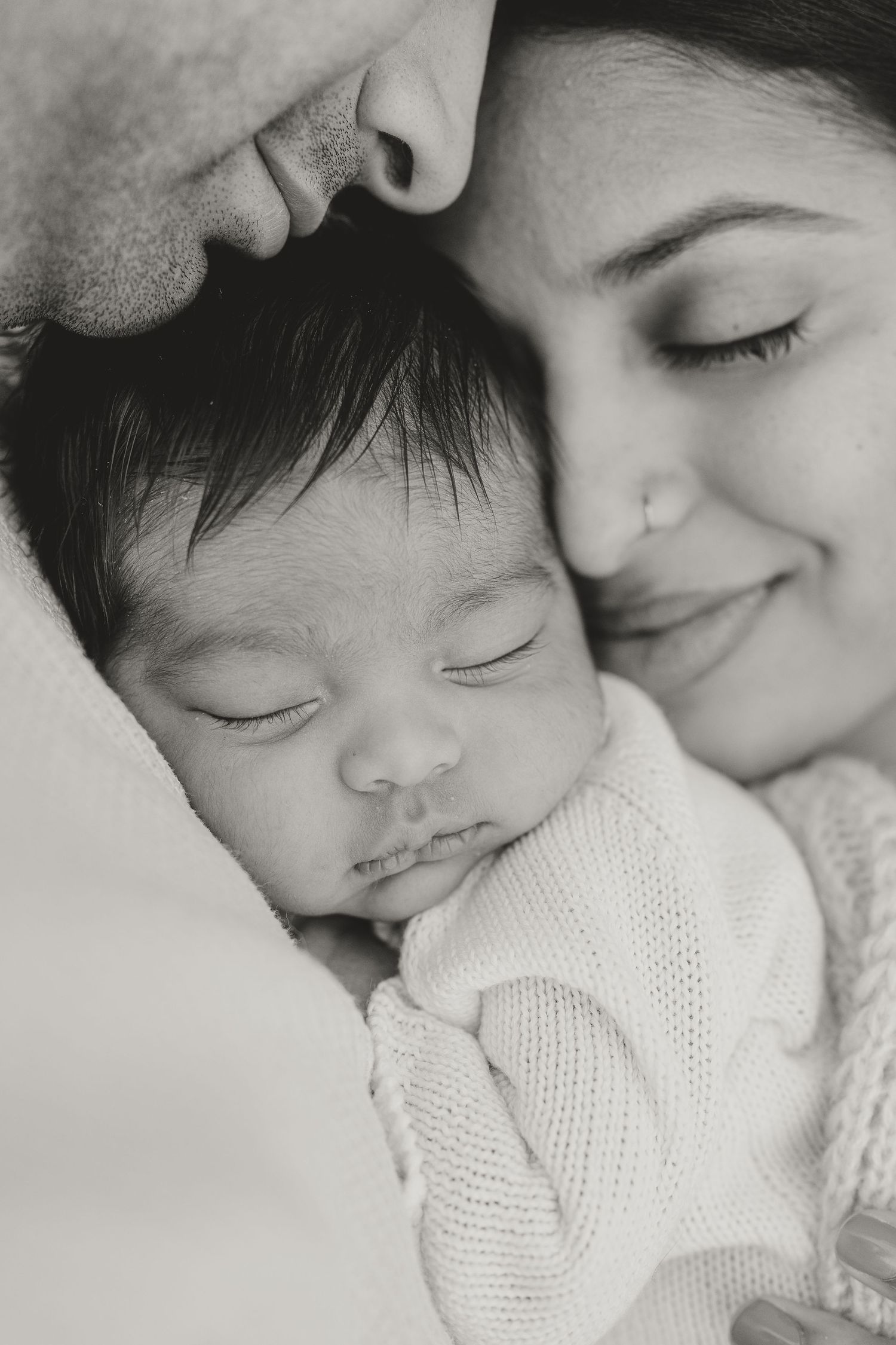 Black and white photo of sleeping newborn baby cradled between smiling parents in a tender family portrait.