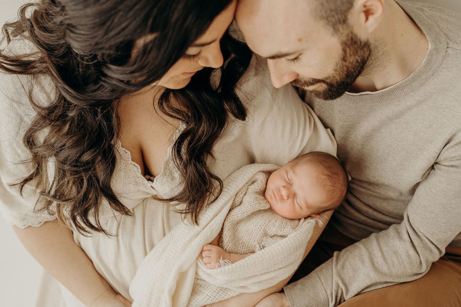 New parents lovingly gaze at their sleeping newborn baby wrapped in a cream swaddle blanket.