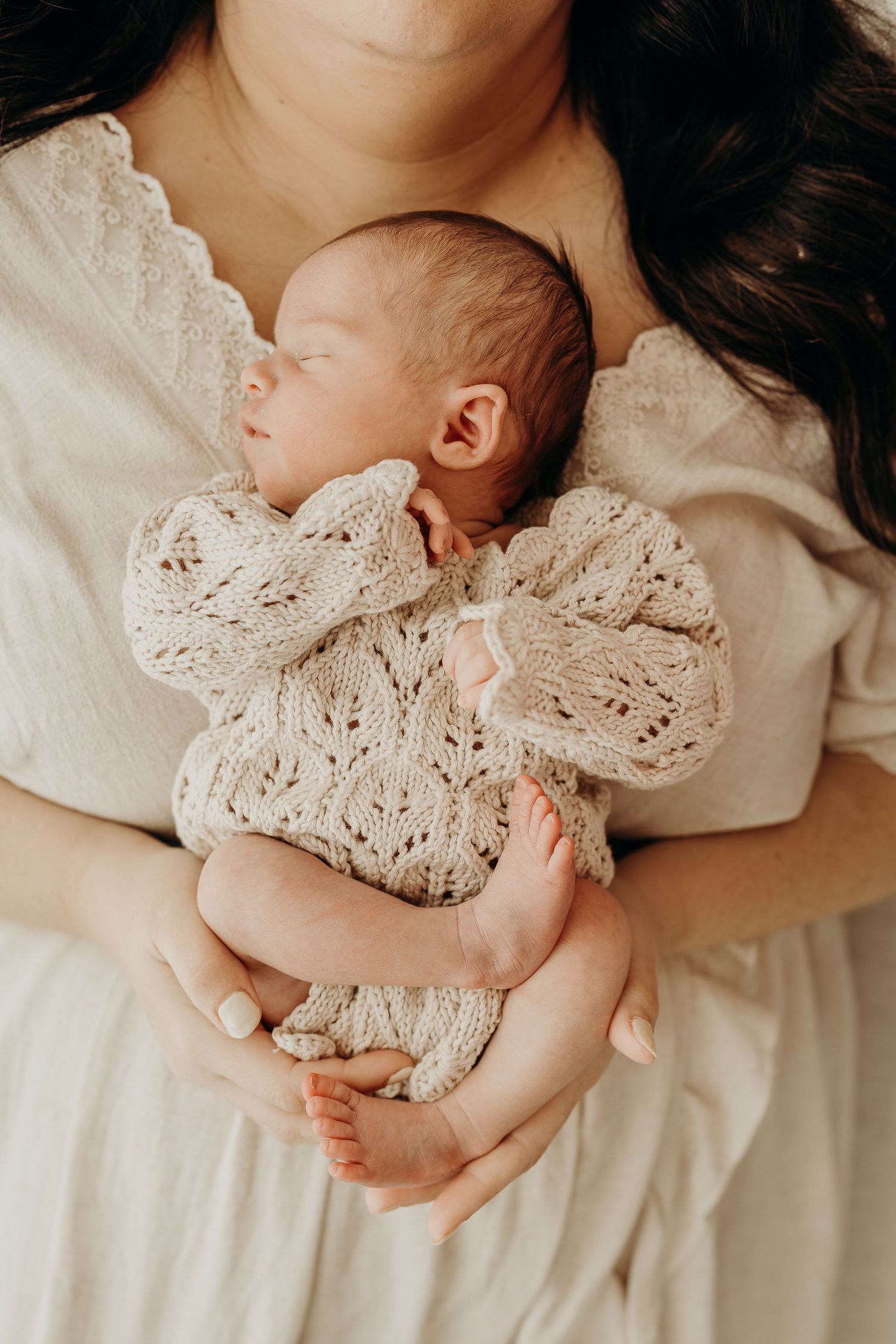Mother in cream dress cradling sleeping newborn baby wrapped in a delicate knit blanket.