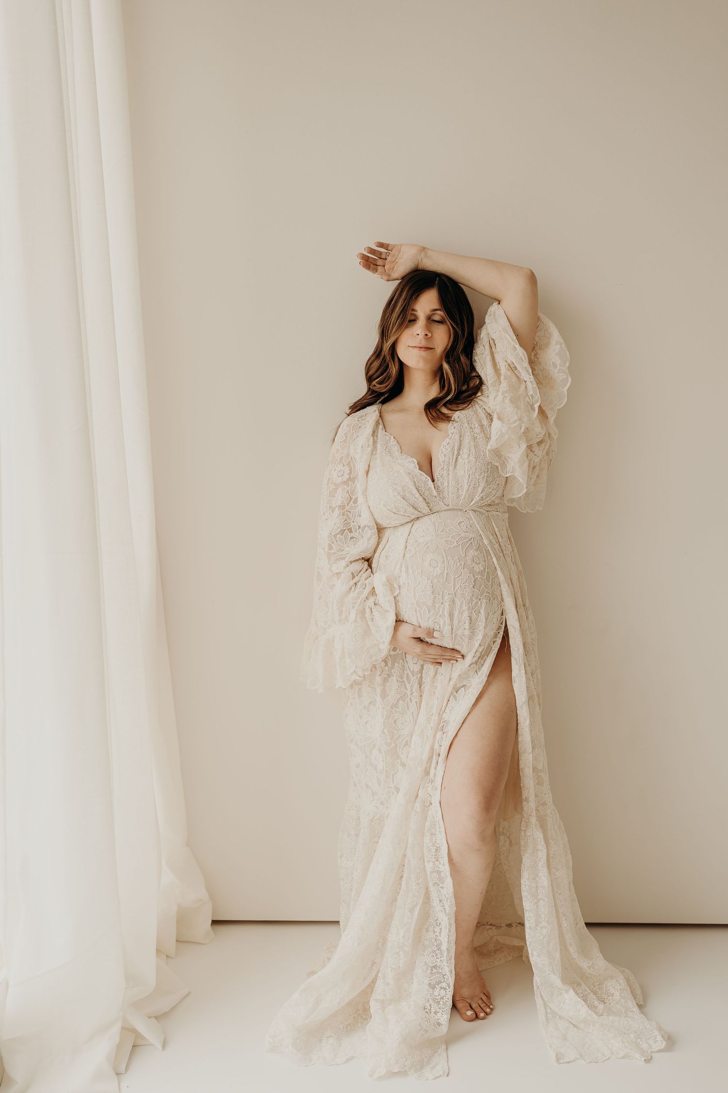 A pregnant woman in a flowing white lace maternity gown poses elegantly against a light backdrop.