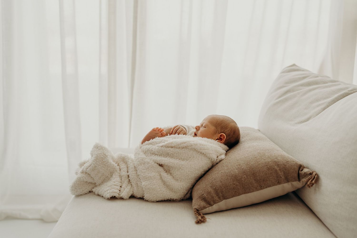 A peaceful newborn baby sleeping wrapped in a white knit blanket on a cream sofa with neutral pillows.