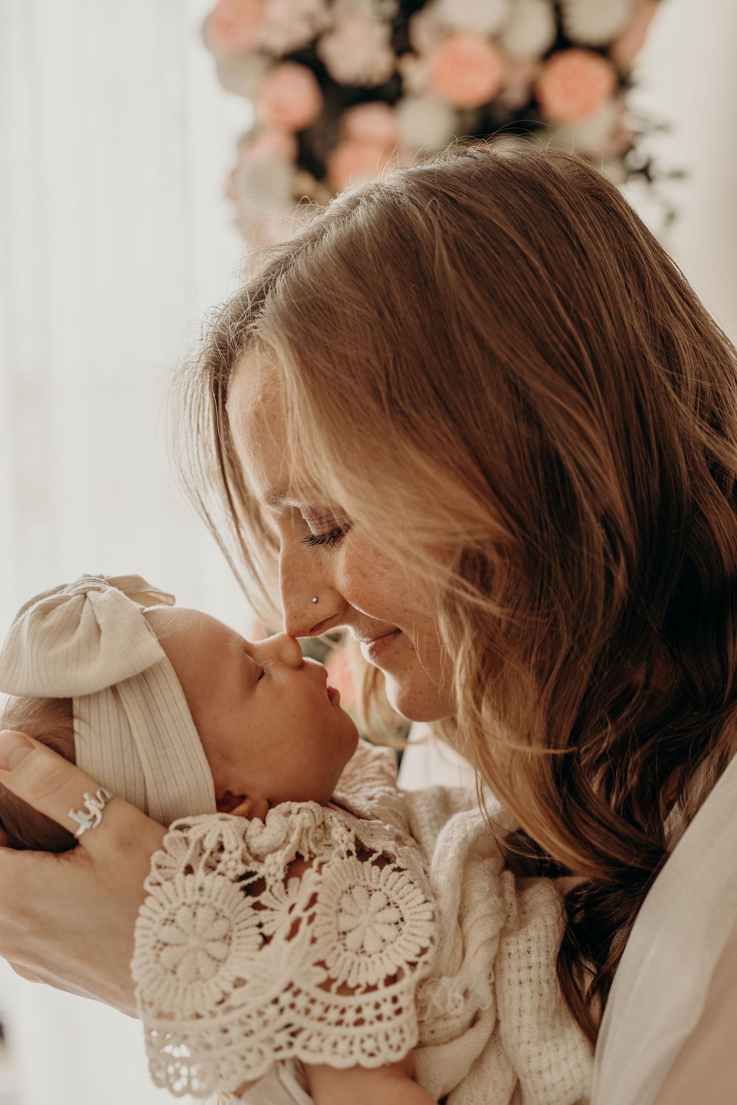 A mother tenderly touches noses with her newborn baby, both dressed in cream lace, with soft floral decor behind them.