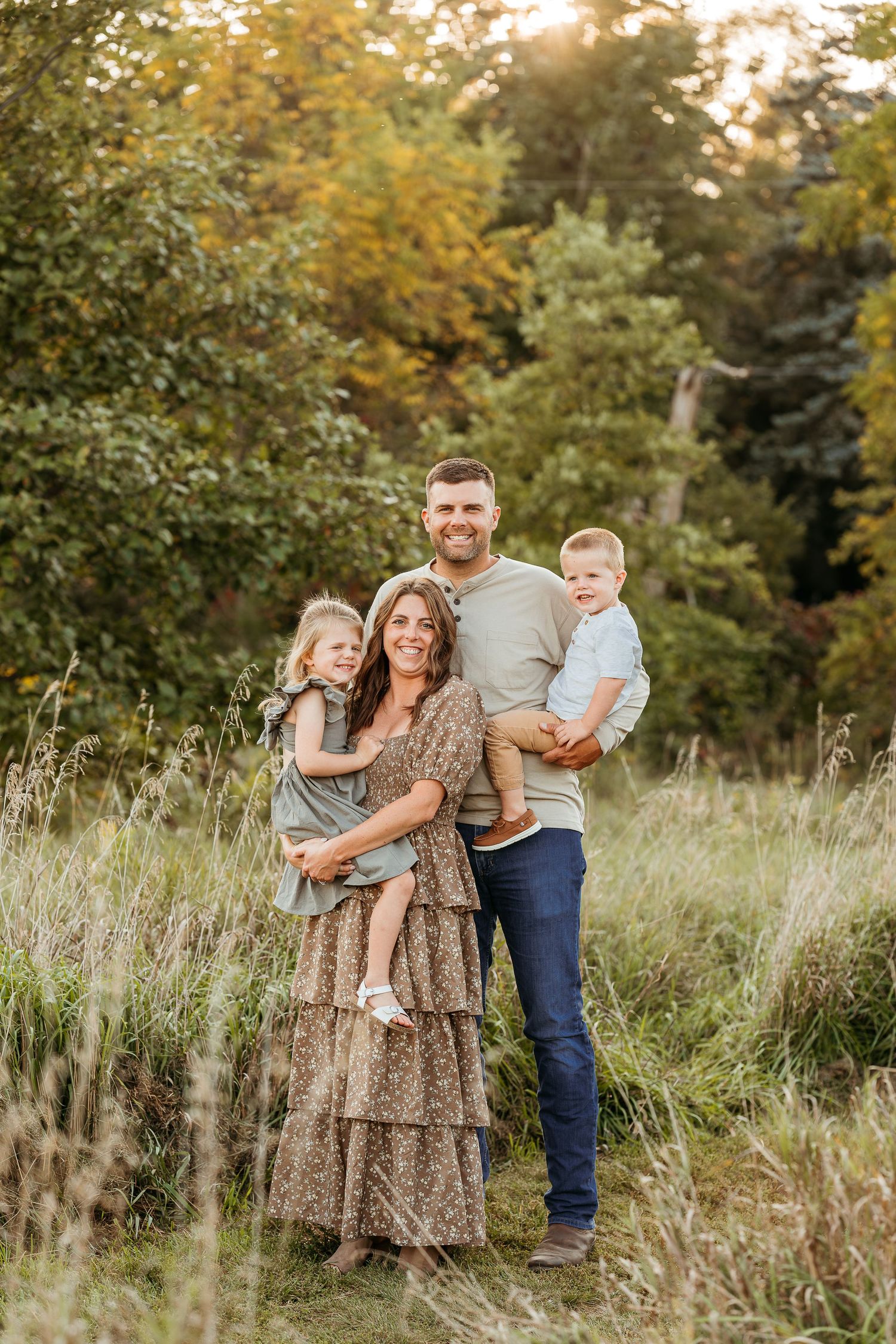 A family poses together in a sunlit meadow with tall grass and autumn trees in the background.