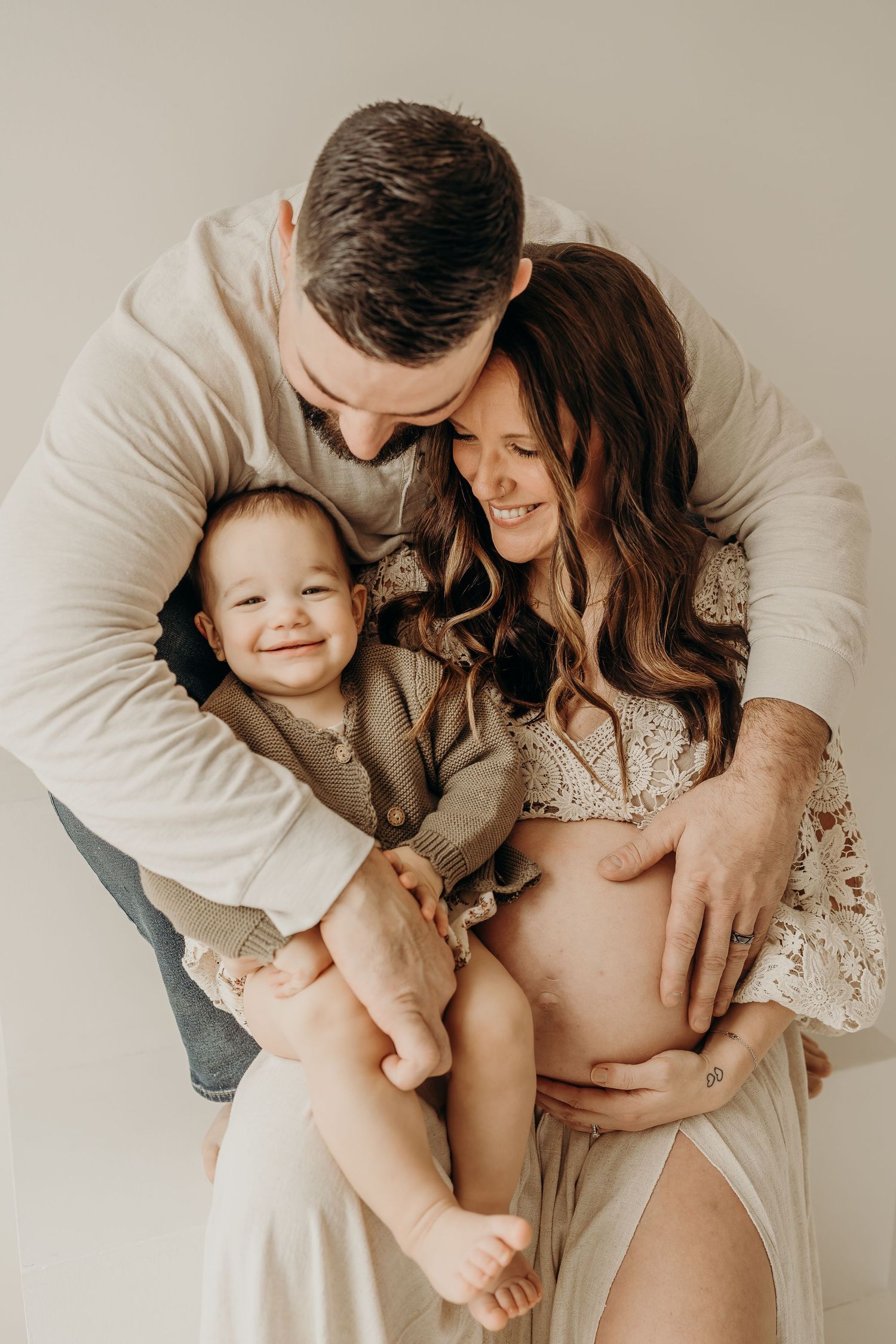 Expecting couple holds smiling toddler during maternity photo session, mother in lace dress cradling baby bump.