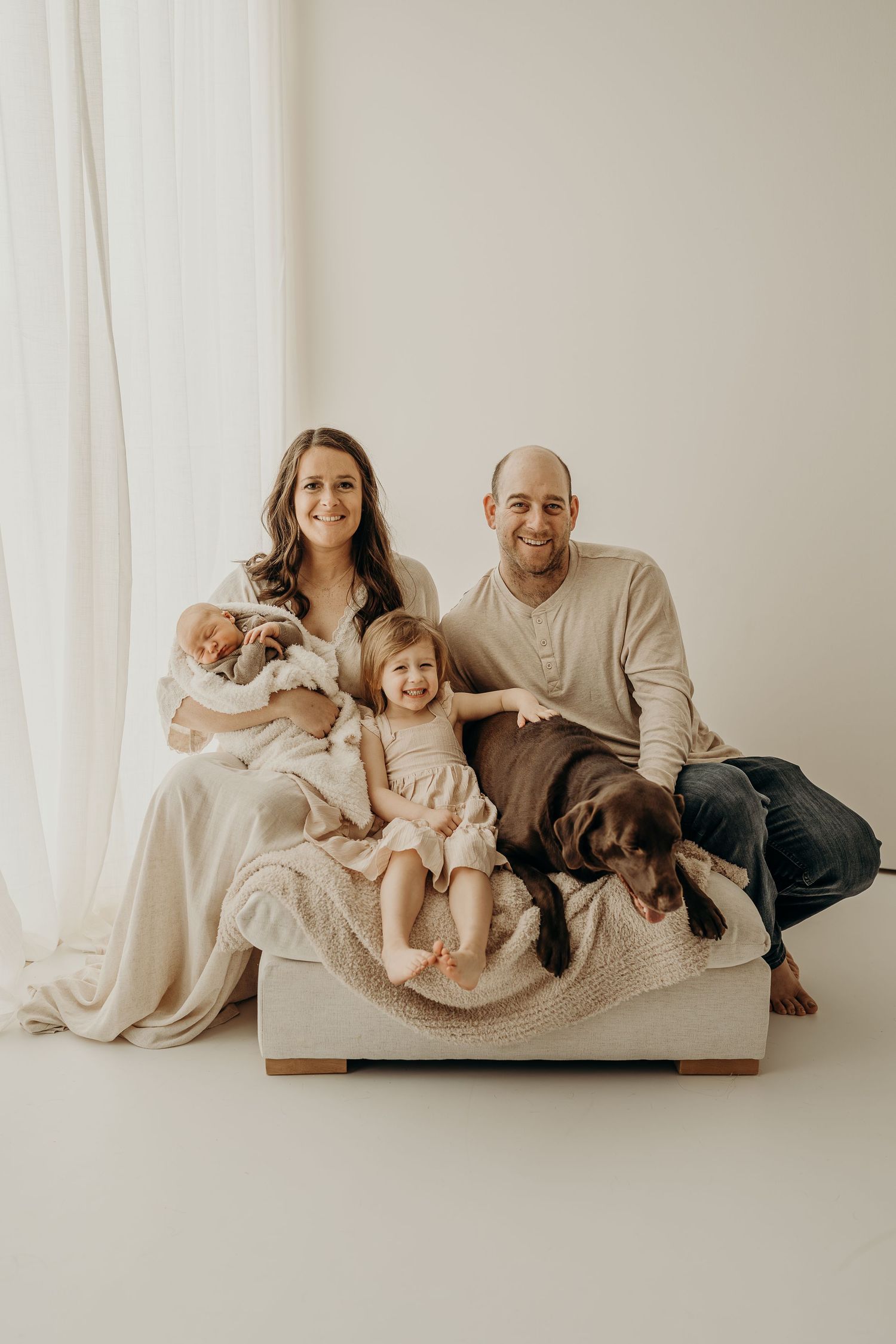 Family portrait with parents, toddler, newborn baby, and brown Labrador dog seated on a couch in a bright studio.