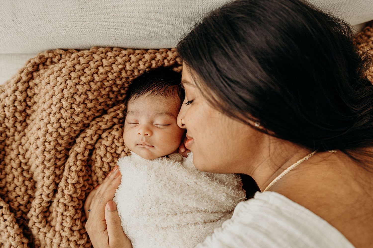A mother tenderly kisses her sleeping newborn baby wrapped in a white blanket on a cozy knit bed.